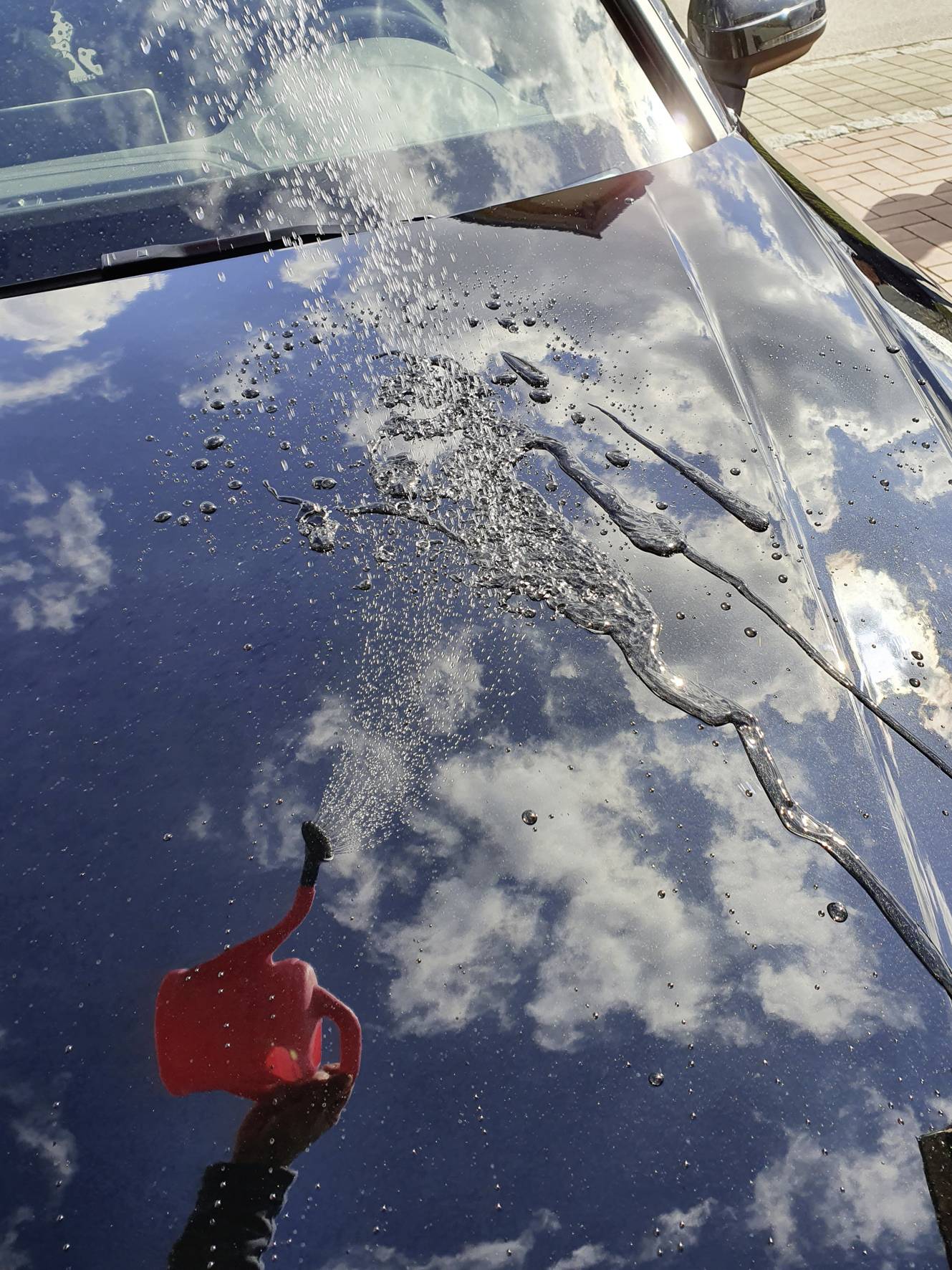 L'eau est versée d'un arrosoir rouge sur le capot brillant d'une voiture, sur lequel se reflètent le ciel et ses nuages.