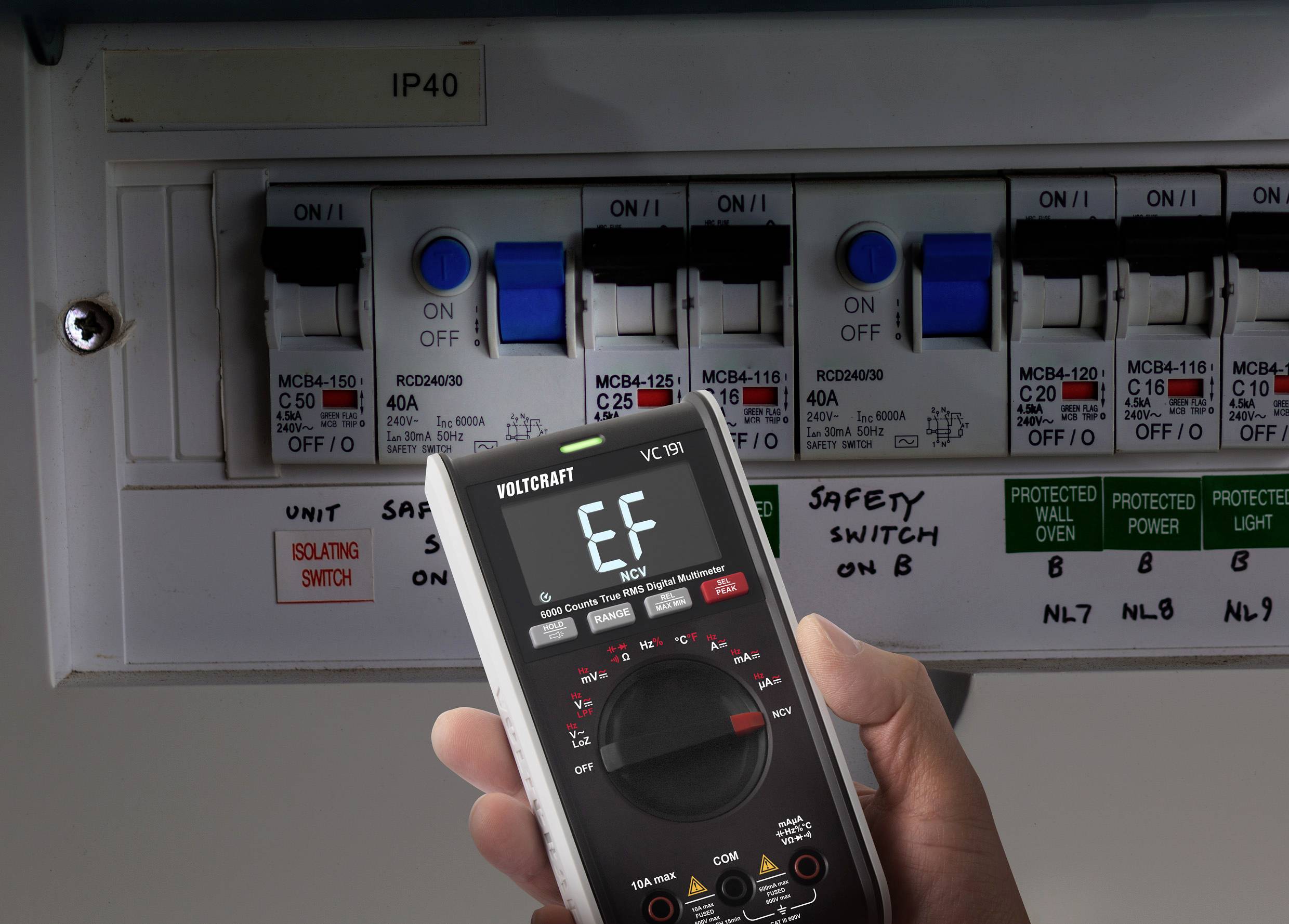 Une personne tient un multimètre numérique affichant une valeur de mesure devant un tableau électrique avec plusieurs disjoncteurs dans un bâtiment.
