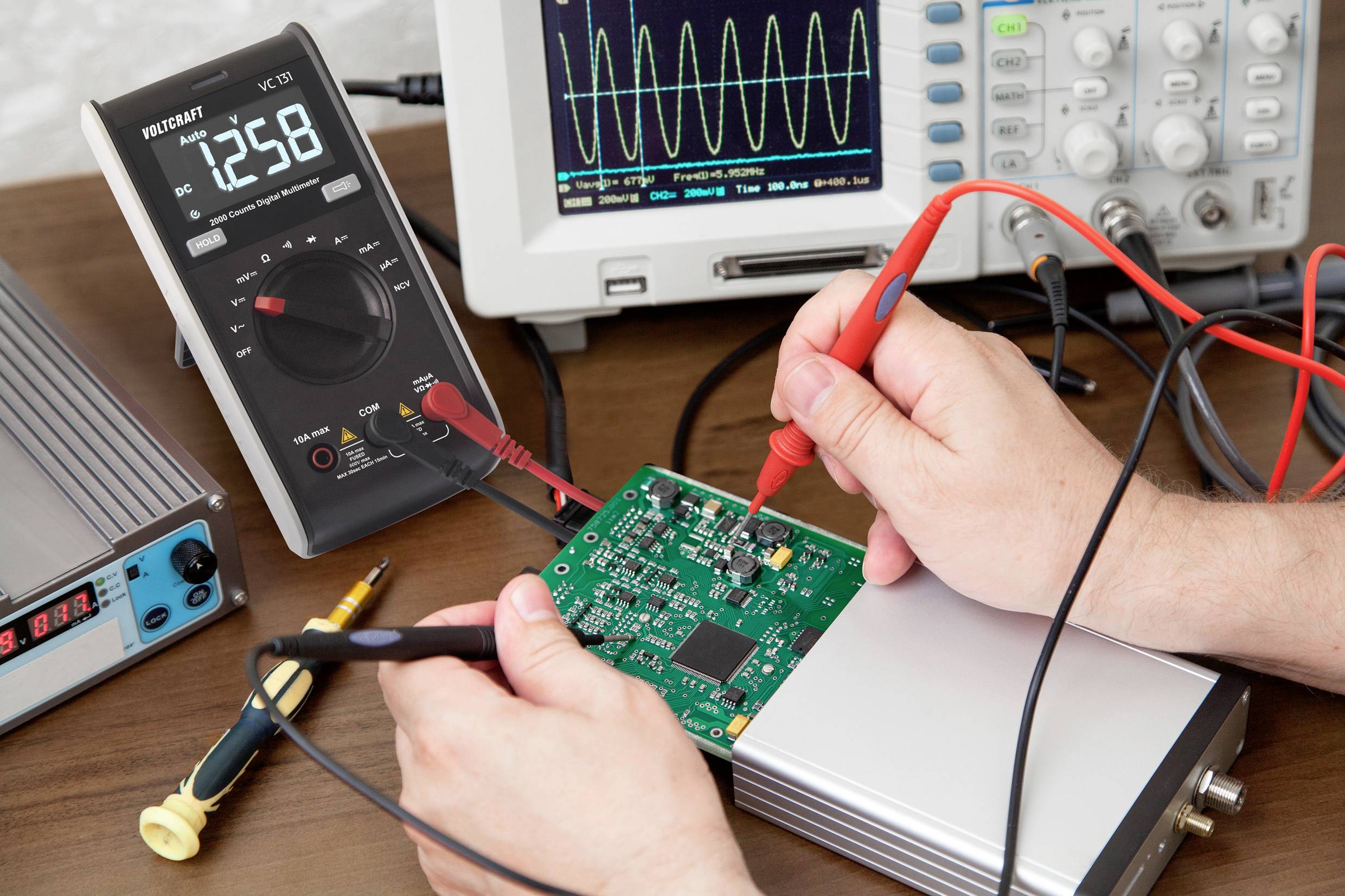 Une personne teste une carte électronique verte avec un multimètre posé sur une table. Un oscilloscope est visible en arrière-plan.