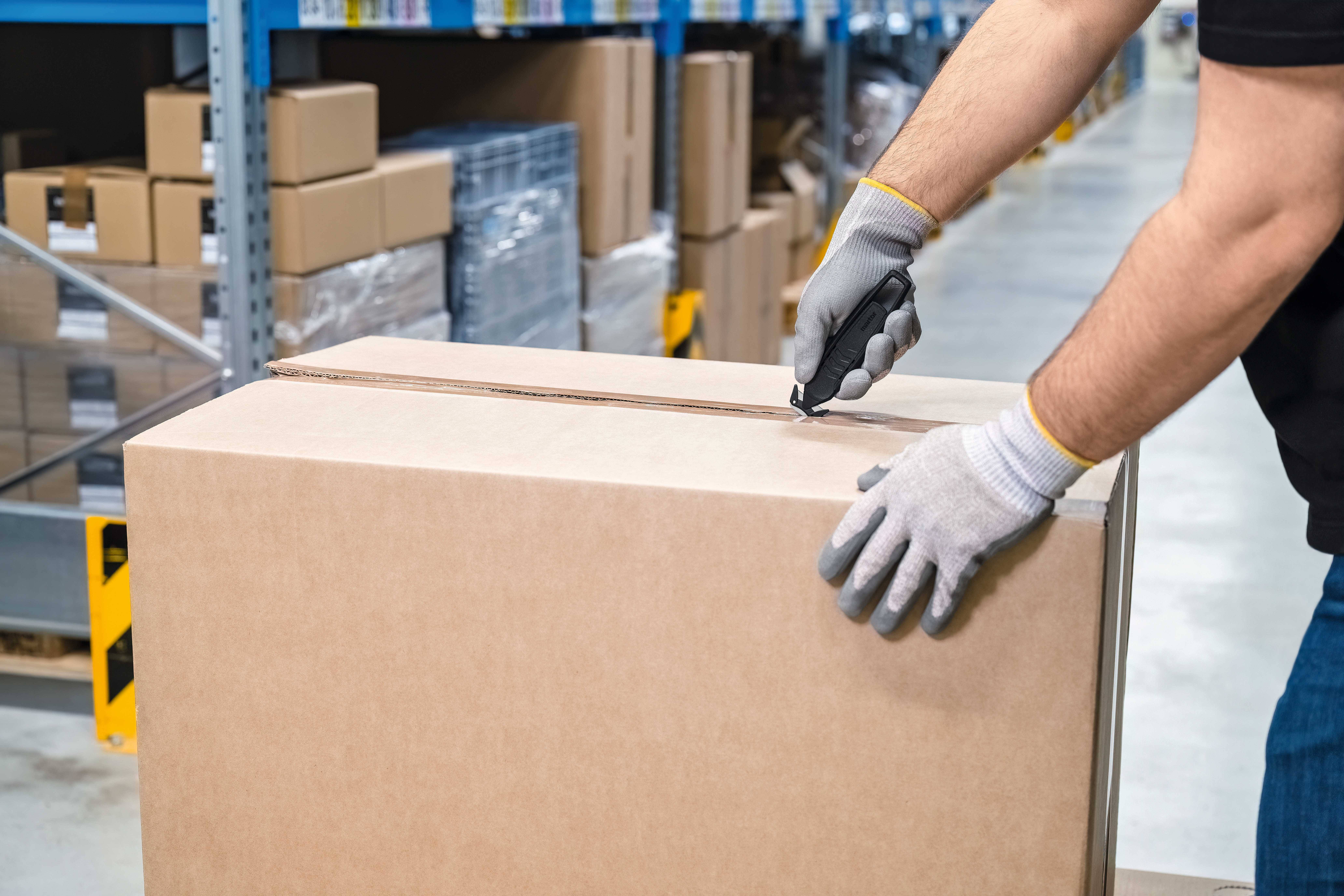 Une personne ouvre un grand carton à l'aide d'un cutter dans un entrepôt, entourée d'étagères remplies d'autres colis et boîtes.