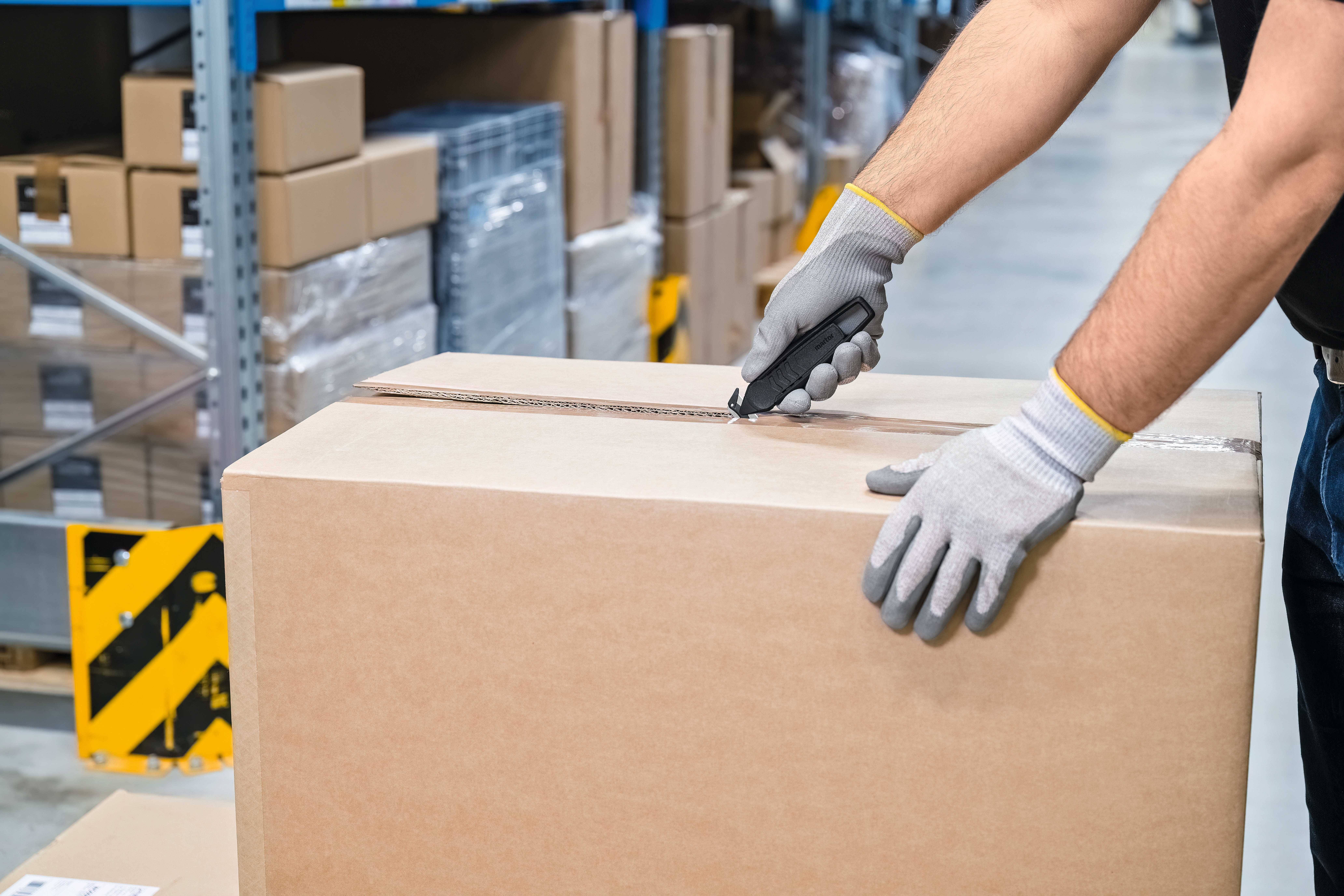 Une personne ouvre un carton avec un couteau utilitaire dans un environnement d'entrepôt. Les mains portent des gants. À l'arrière-plan, on peut voir des étagères avec des marchandises.