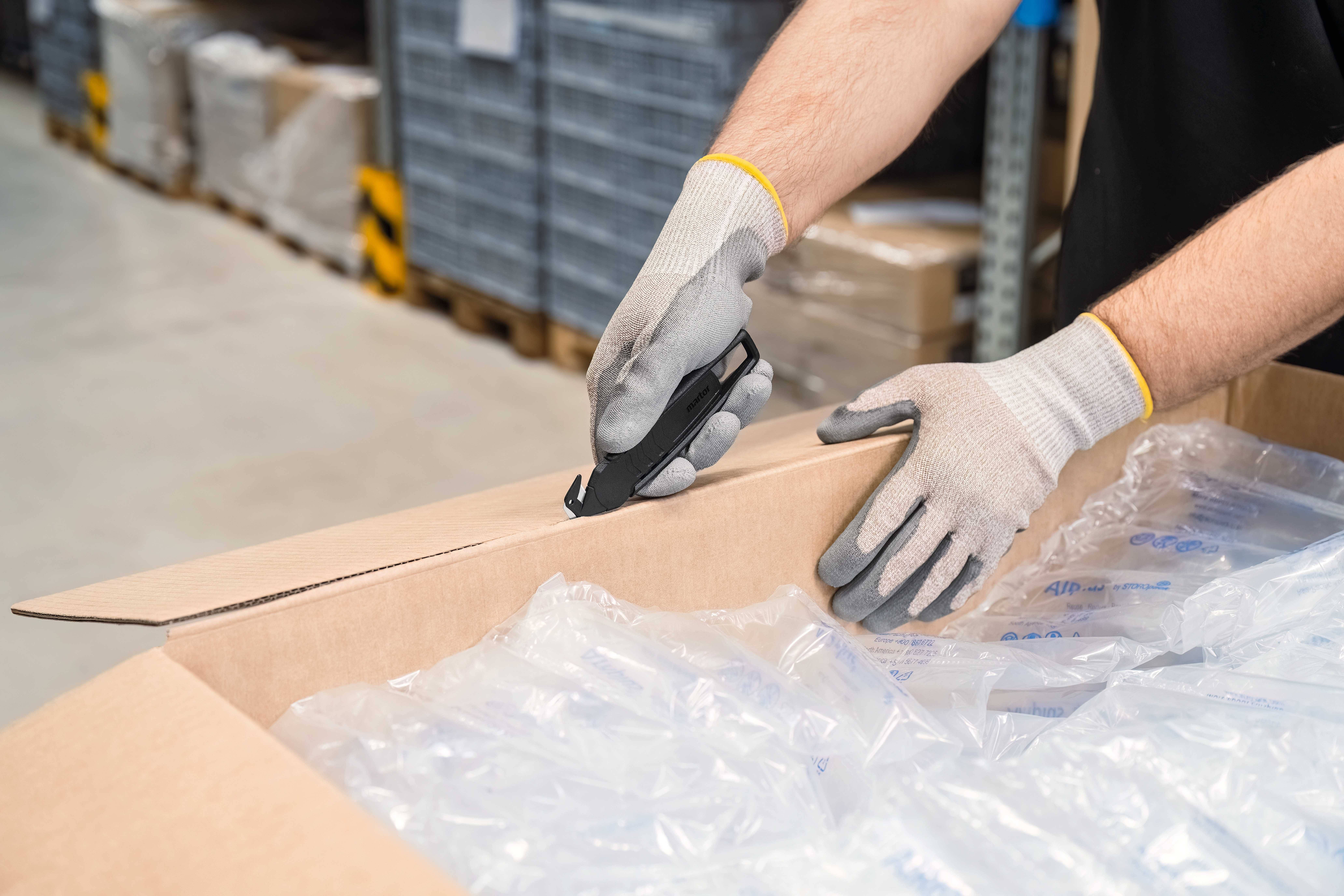 Une personne portant des gants ouvre un carton à l'aide d'un couteau dans un entrepôt, rempli de film à bulles.