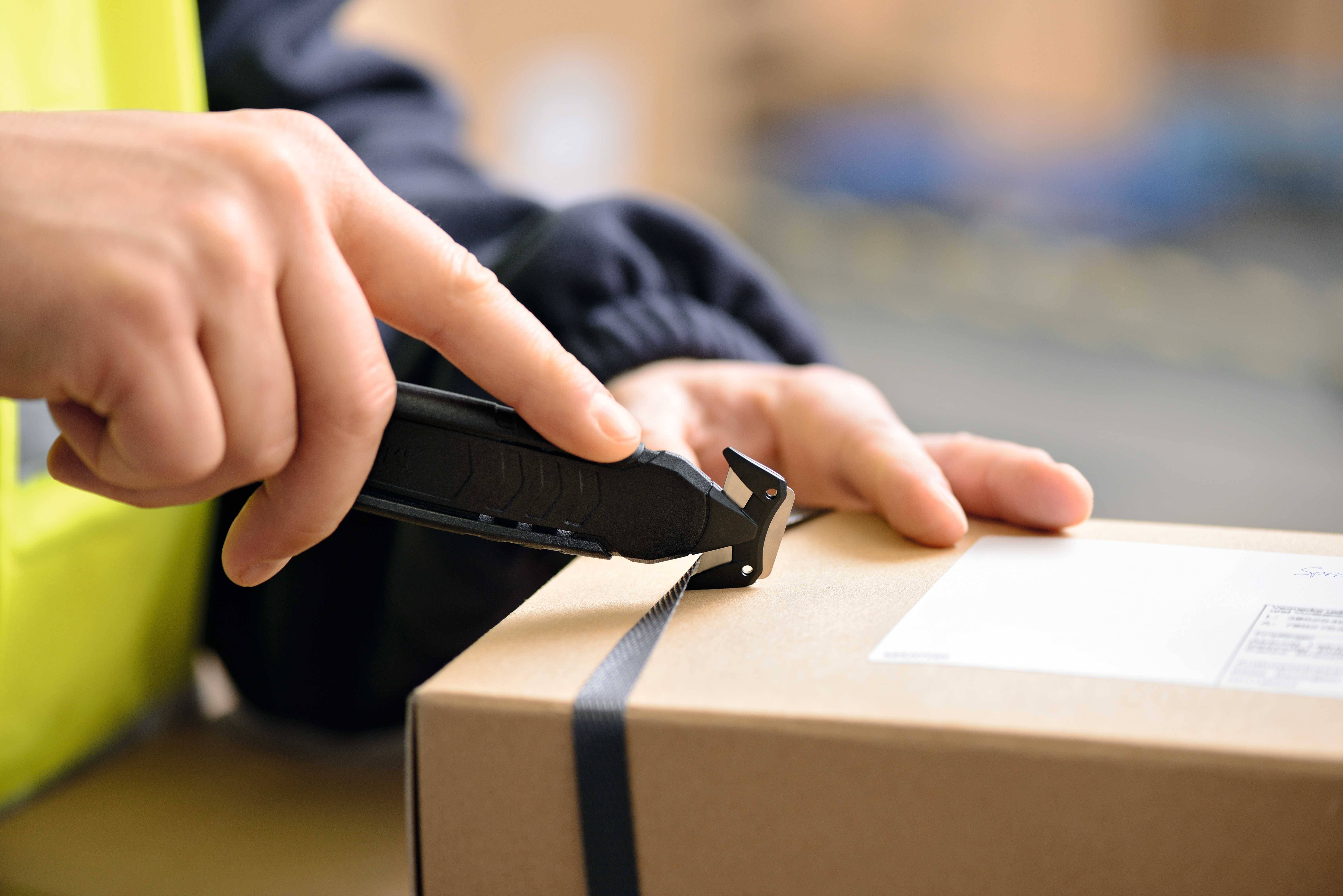 Une personne ouvre prudemment un carton scellé avec du ruban adhésif noir à l'aide d'un cutter.