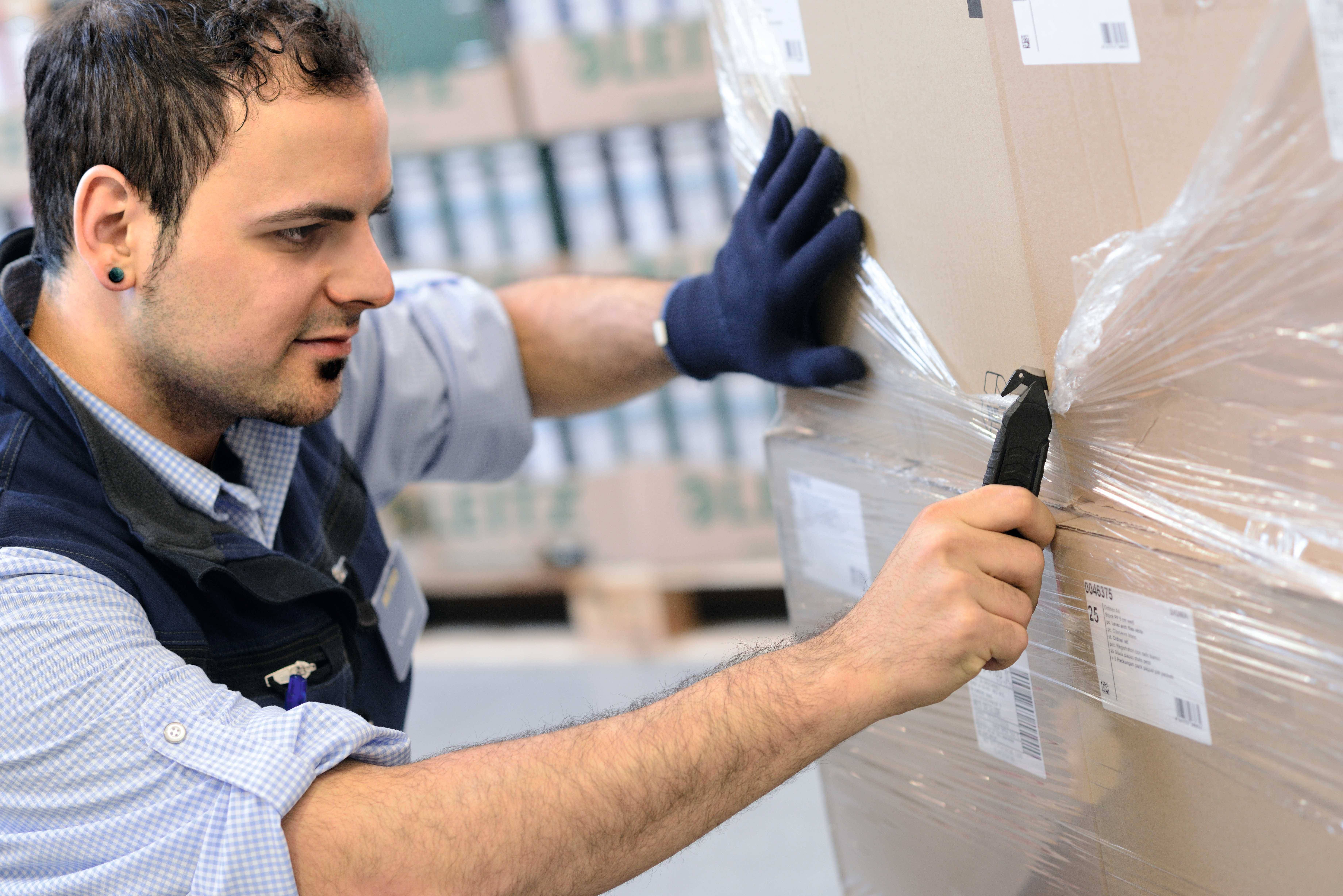 Un homme portant des gants et tenant un couteau ouvre une caisse enveloppée de film plastique dans un entrepôt.