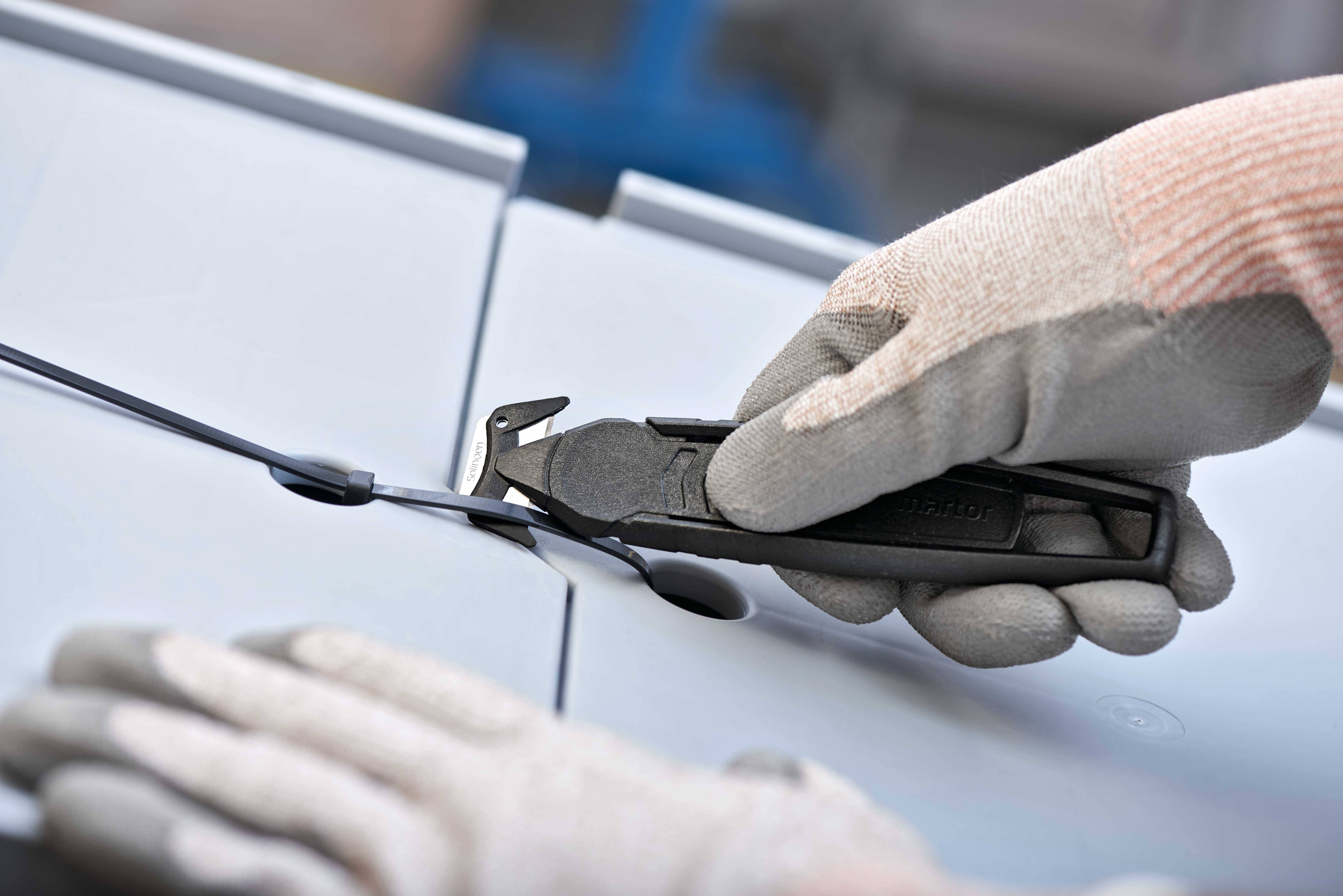 Une main gantée coupe un câble avec un coupe-fil sur un fond gris.