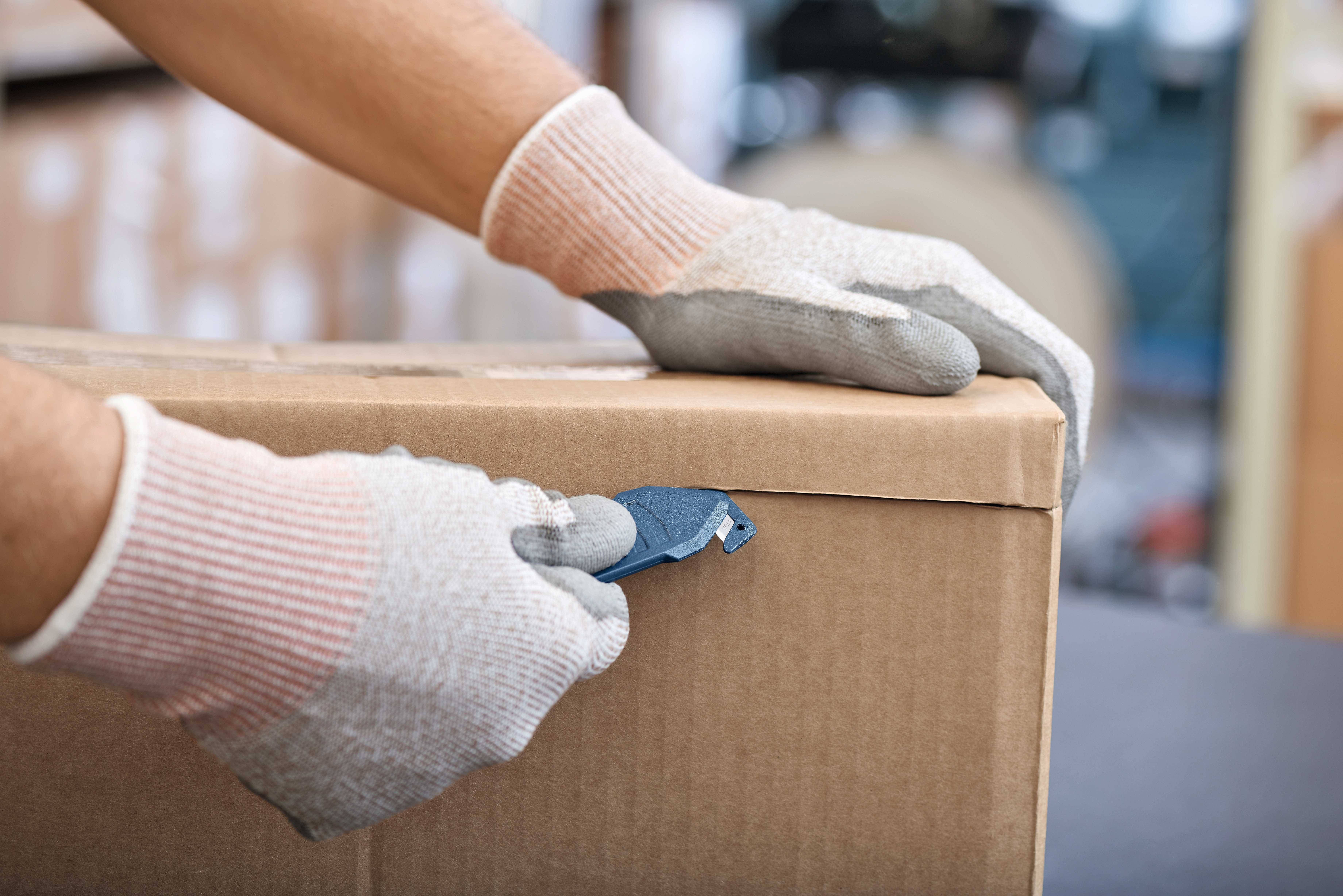 Une personne ouvre un carton en carton à l'aide d'un cutter. Les mains sont protégées par des gants de travail.