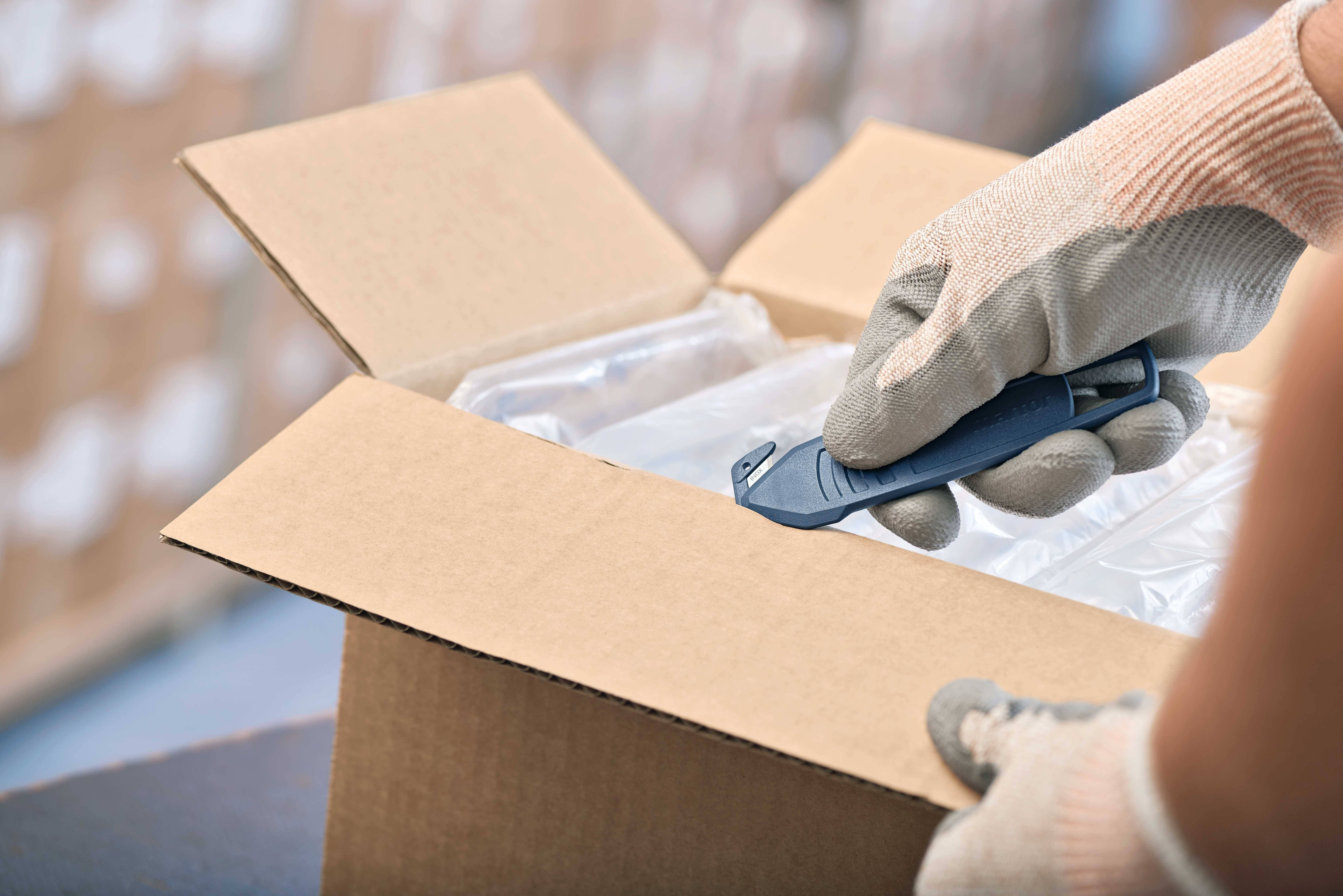 Une personne portant des gants ouvre un carton avec un cutter. Des étagères d'entrepôt floues sont visibles en arrière-plan.