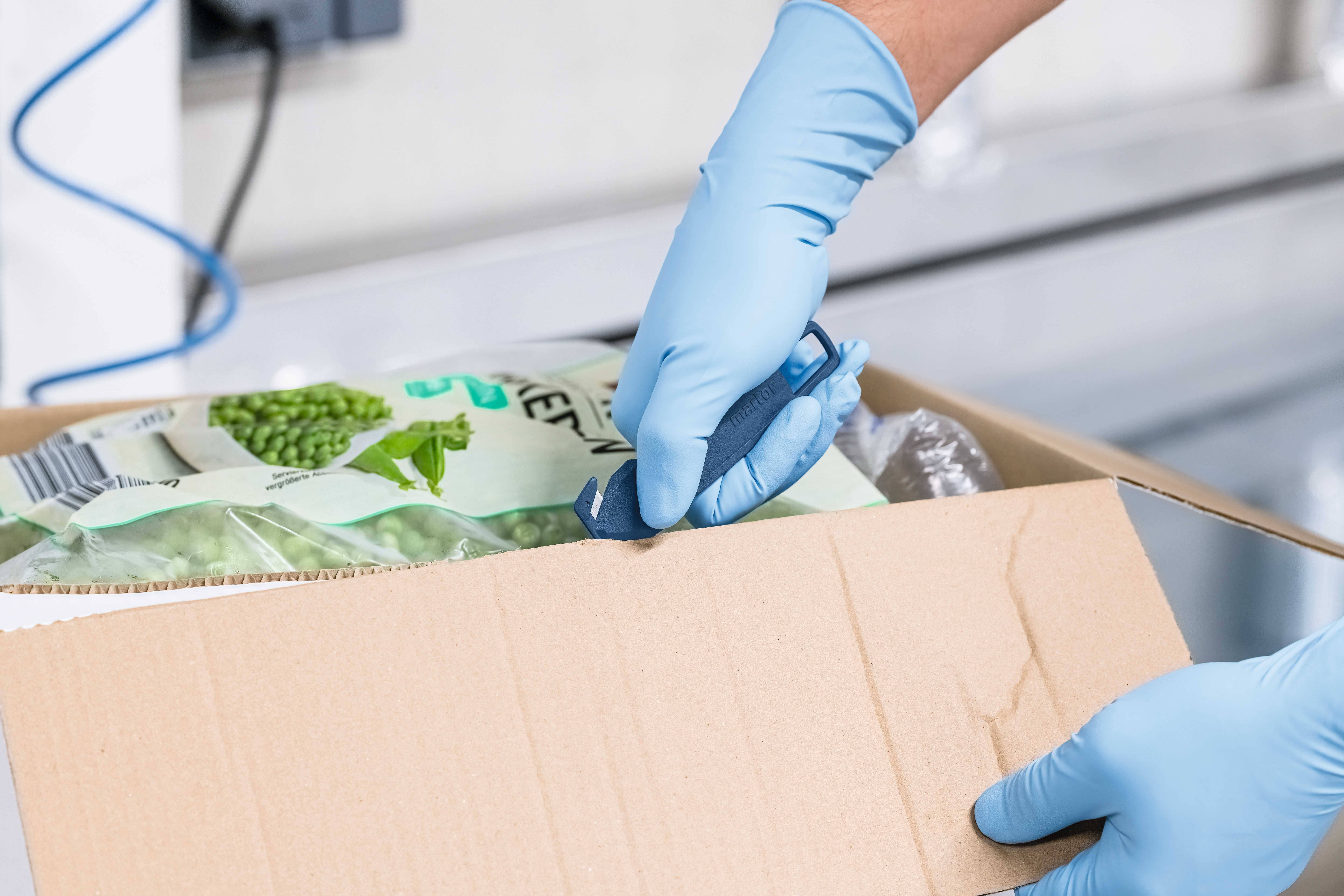 Une personne ouvre une boîte en carton à l'aide d'un petit couteau. Elle porte des gants bleus. Dans la boîte se trouvent des petits pois surgelés.