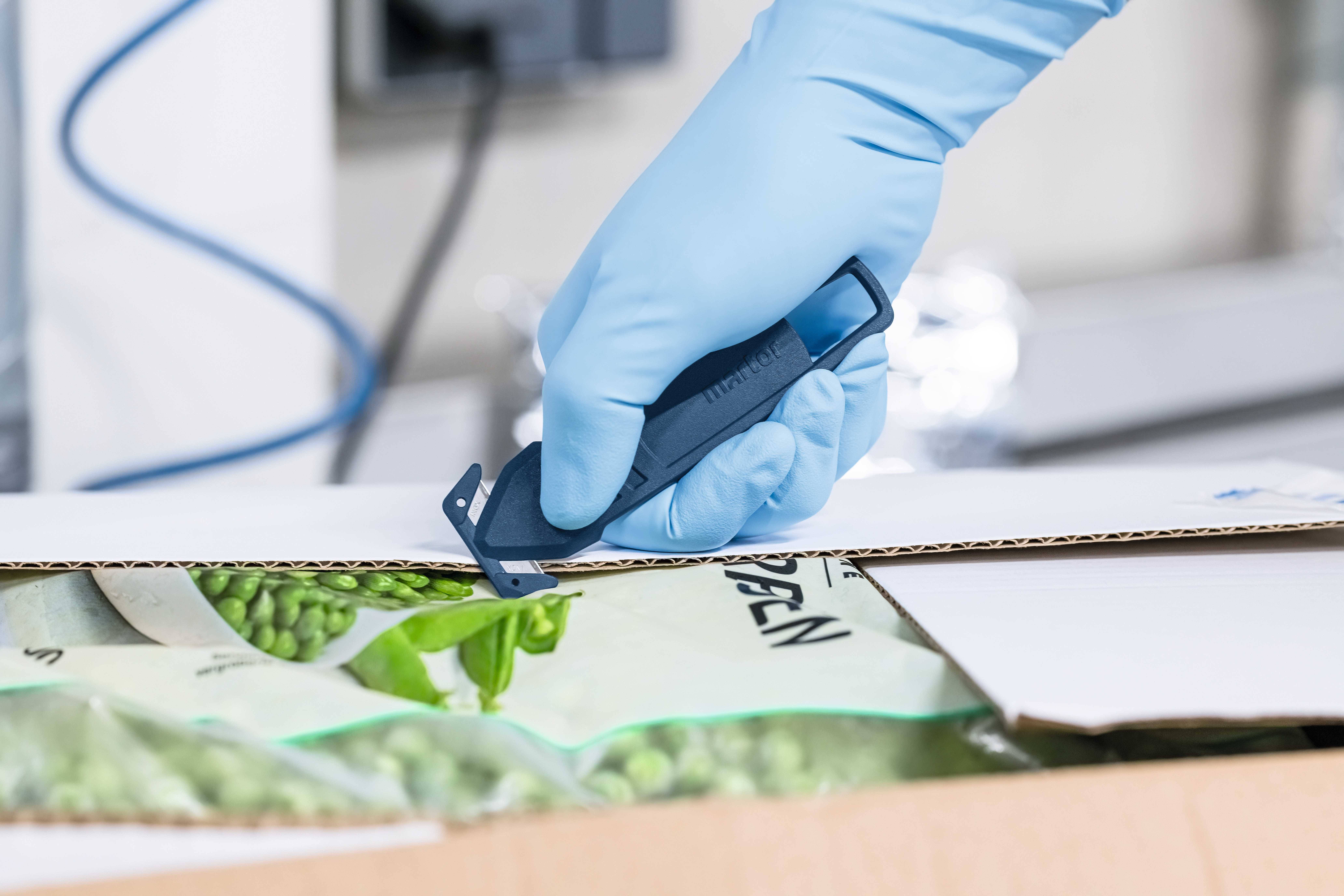 Une main gantée d'un gant bleu ouvre un carton contenant des légumes surgelés à l'aide d'un couteau.