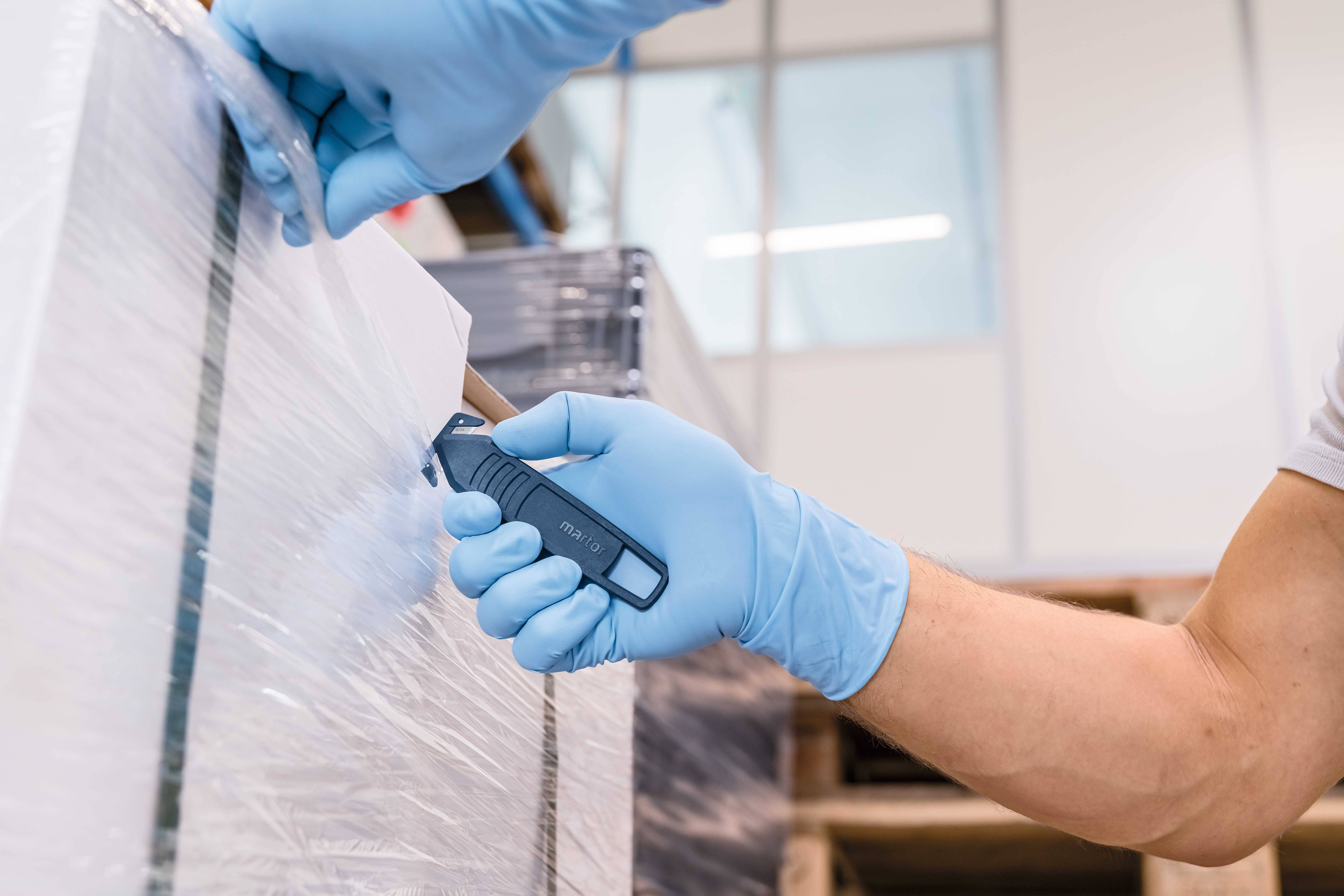 Une personne portant des gants bleus coupe un film plastique d'une palette à l'aide d'un couteau. Un entrepôt est visible en arrière-plan.