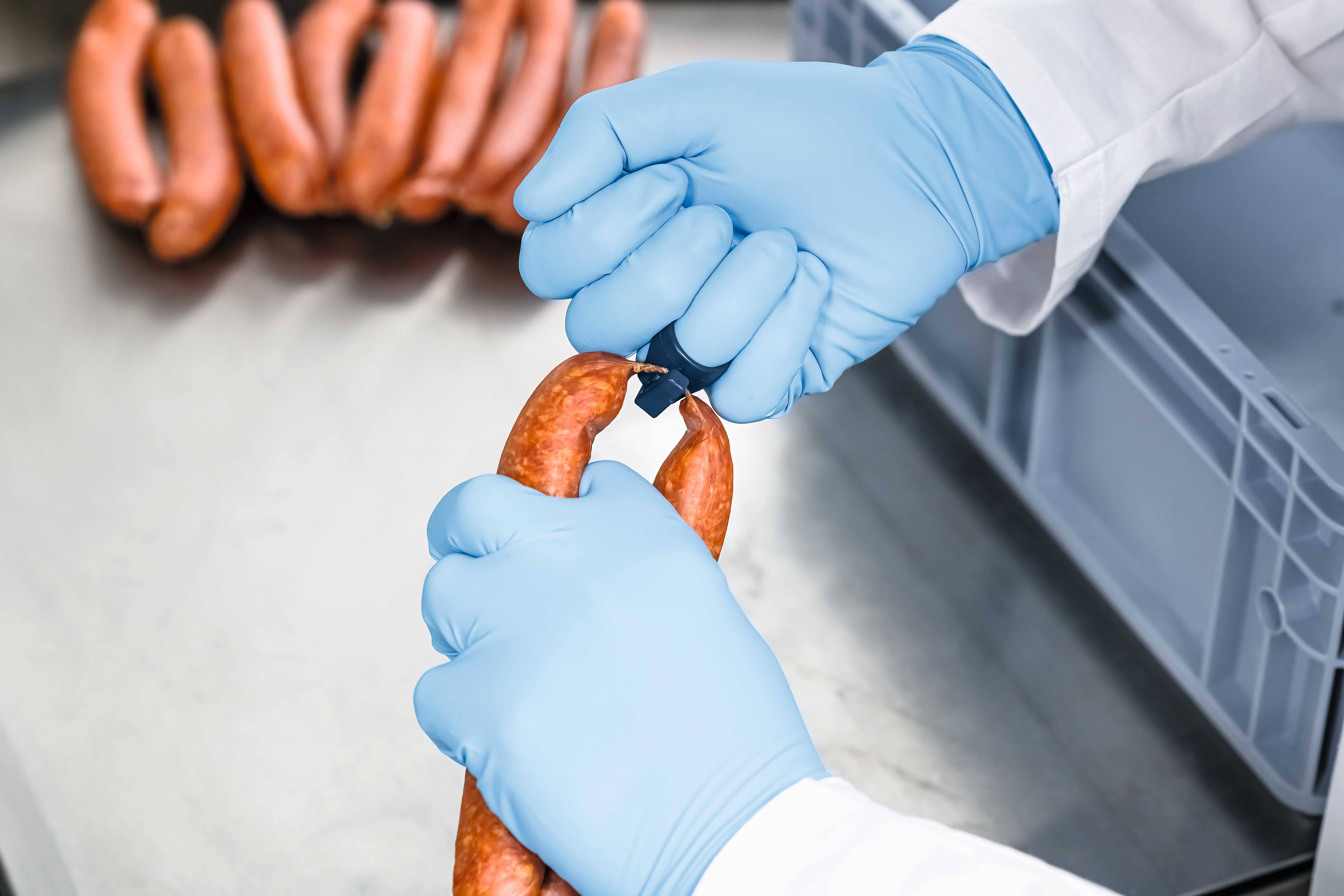 Une personne portant des gants bleus attache les extrémités d'une saucisse avec un lien. À l'arrière-plan, plusieurs saucisses sont visibles sur une table.