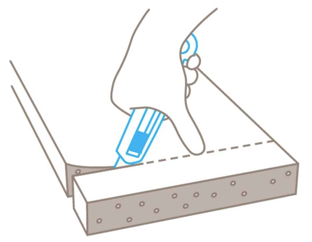 Une main tient un cutter bleu et coupe le long d'une ligne pointillée à travers un morceau de matériau.