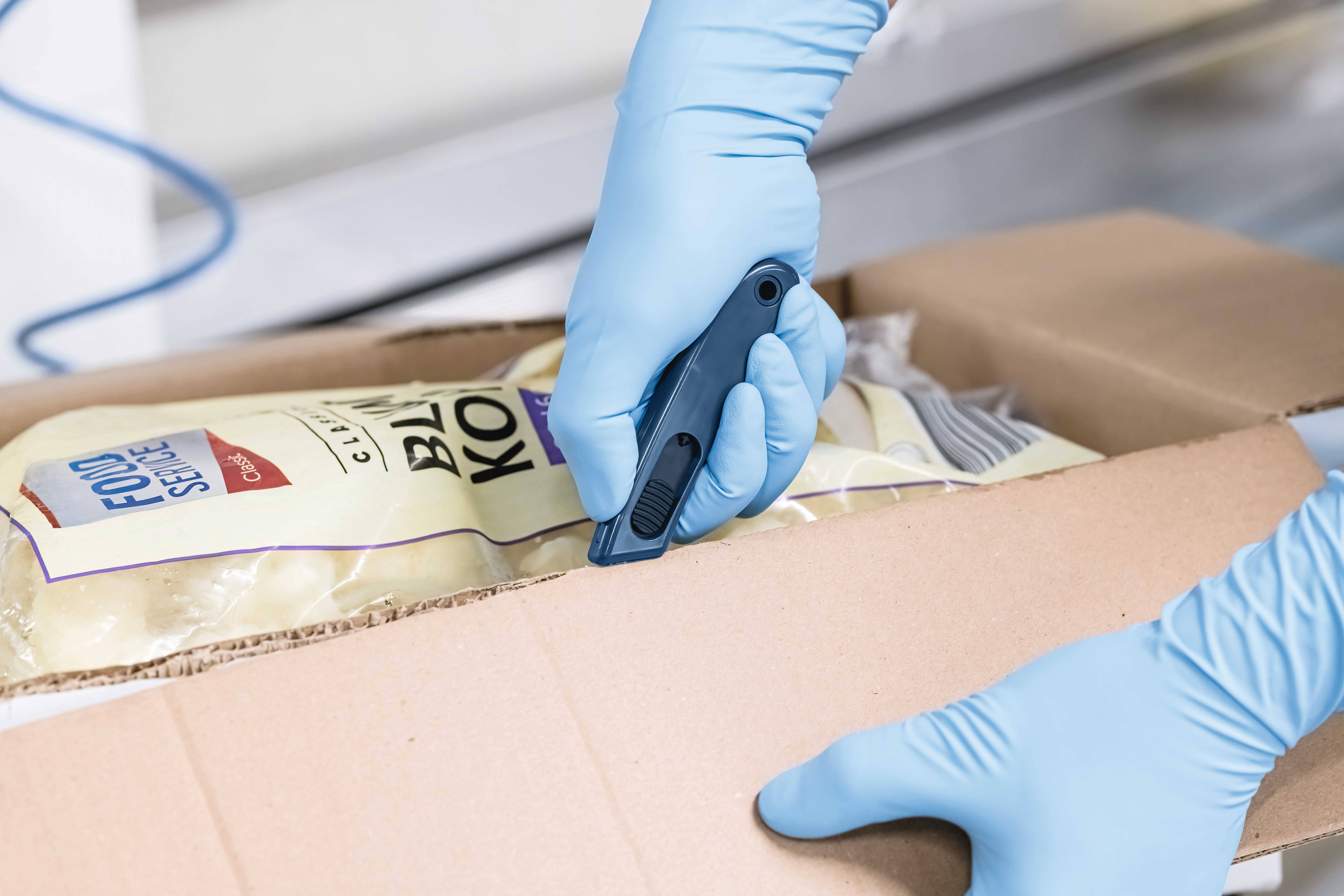 Une personne ouvre prudemment un carton à l'aide d'un couteau de sécurité. À l'intérieur du carton se trouvent des produits sous vide, identifiables grâce à leurs étiquettes.