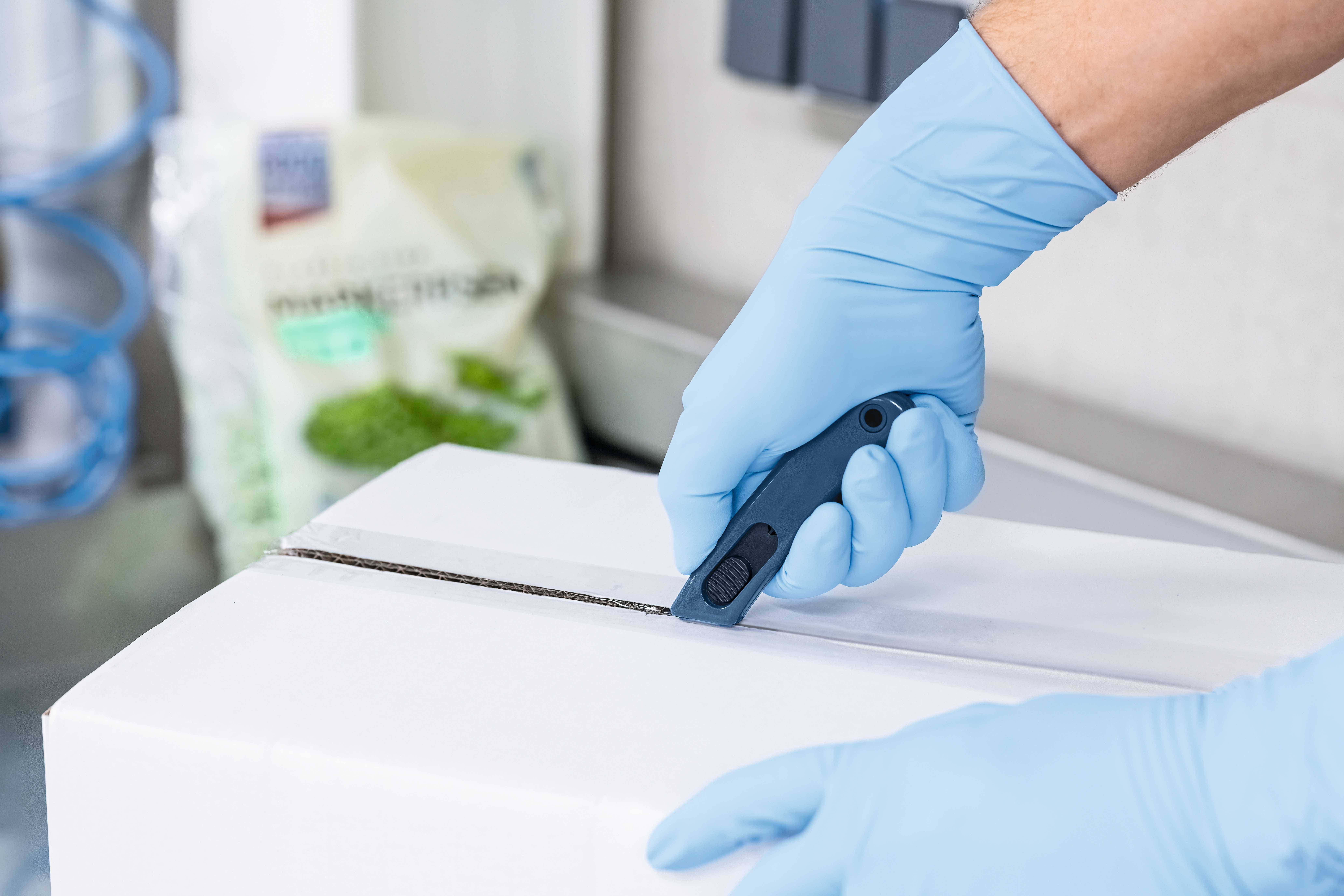 Une personne coupe un paquet blanc avec un cutter noir. Elle porte des gants jetables bleus. À l'arrière-plan, on distingue un sac contenant un contenu vert.