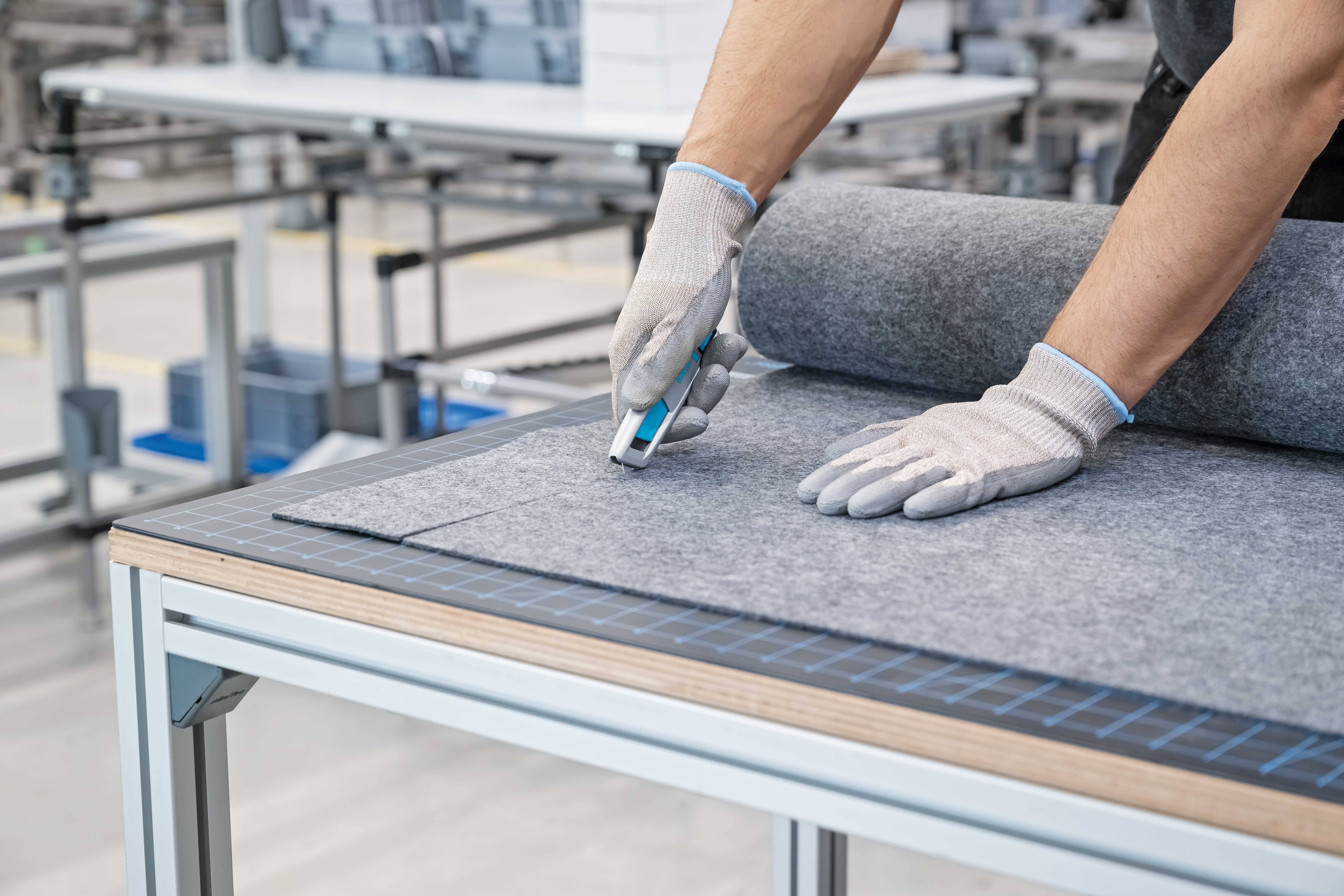Un ouvrier portant des gants coupe du tissu gris avec un couteau utilitaire sur une table dans une usine.