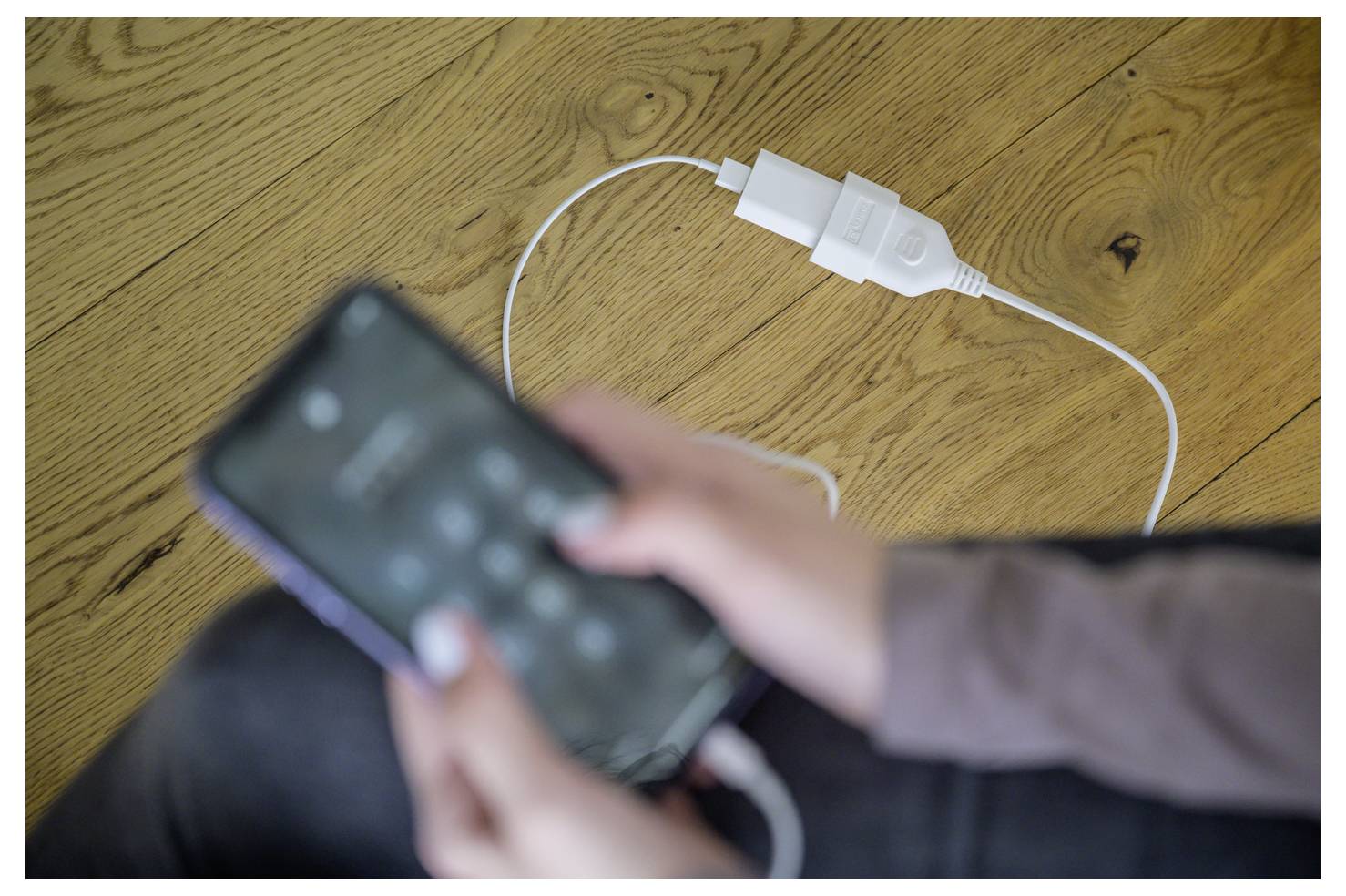 Personne floue utilisant un téléphone intelligent connecté par un câble de recharge USB à un adaptateur sur un plancher en bois.