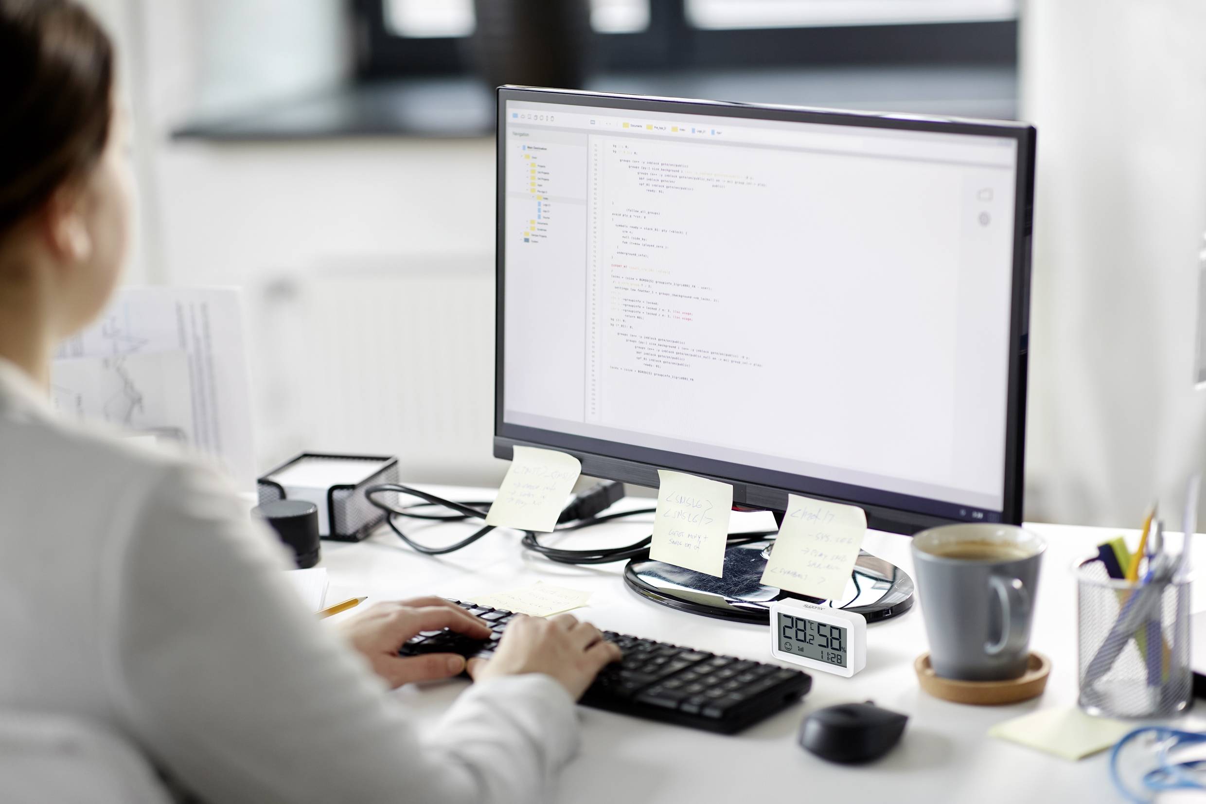 Une personne travaille à un bureau devant un écran affichant du code de programmation. Le bureau est équipé d'une tasse et de notes.