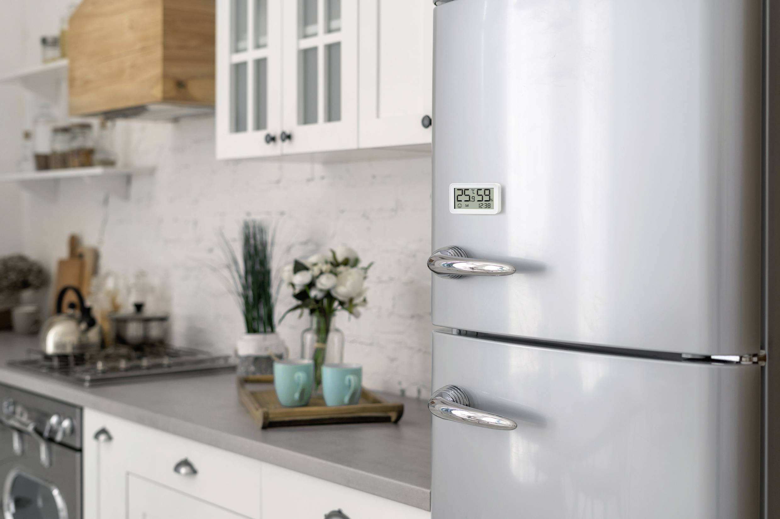 Une cuisine moderne avec un réfrigérateur blanc au premier plan. L'affichage indique '23:58'. À gauche, on peut voir des placards, une cuisinière et une bouilloire.