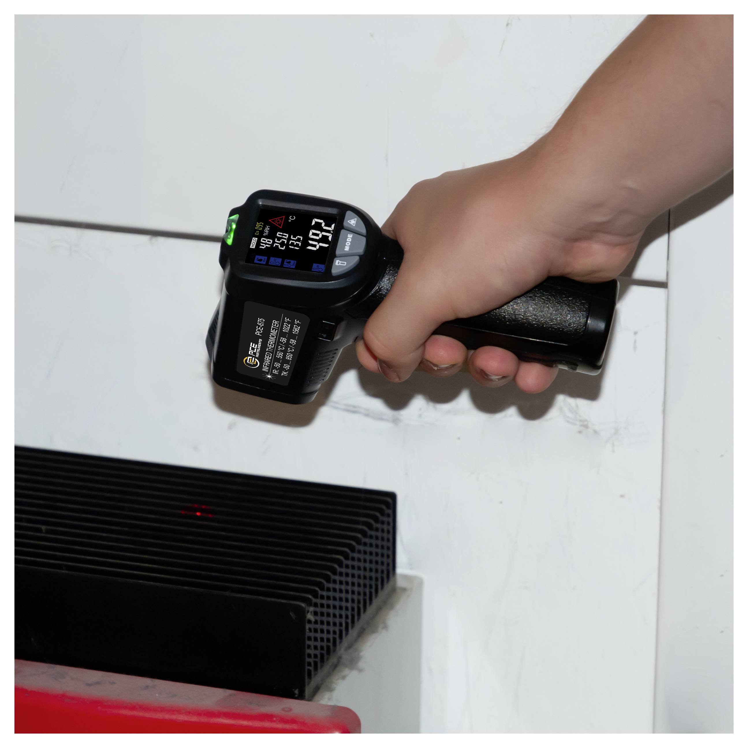 Tragbares Infrarot-Thermometer, das ein schwarzes elektronisches Gerät oder eine Lüftungsöffnung auf einer weißen Oberfläche misst und einen Temperaturwert anzeigt.