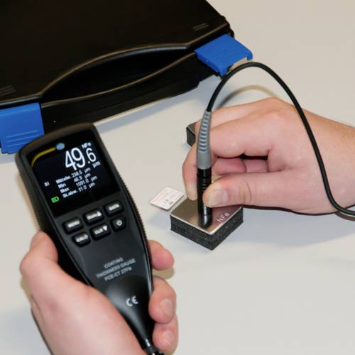 Deux mains manipulent un appareil de mesure. Une main tient l'appareil qui affiche une valeur, tandis que l'autre main dirige une sonde vers un objet de test.