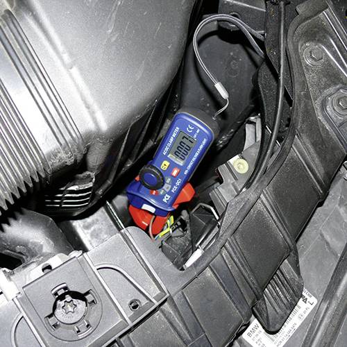 Un voltmètre affiche 12,4 volts et est connecté à une batterie de voiture. L'environnement montre des parties d'un compartiment moteur.