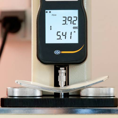 Un dispositif de mesure avec affichage numérique indique 'PEAK 3.92' et '5.41 N'. Il mesure la pression exercée sur un objet légèrement incurvé placé entre deux disques.