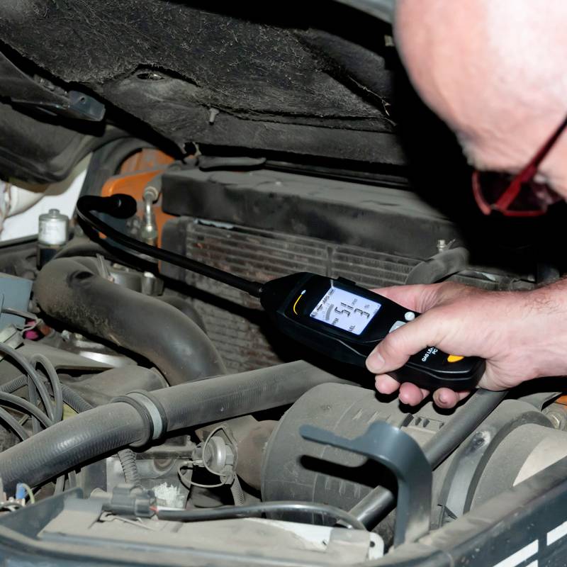 Une personne mesure la température d'un moteur à l'aide d'un thermomètre numérique. Le compartiment moteur est ouvert et expose les pièces du moteur.
