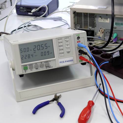 Un appareil de mesure numérique affiche '20,55' sur '220 V AC'. À côté se trouvent un tournevis et une pince. Contexte : environnement de laboratoire.