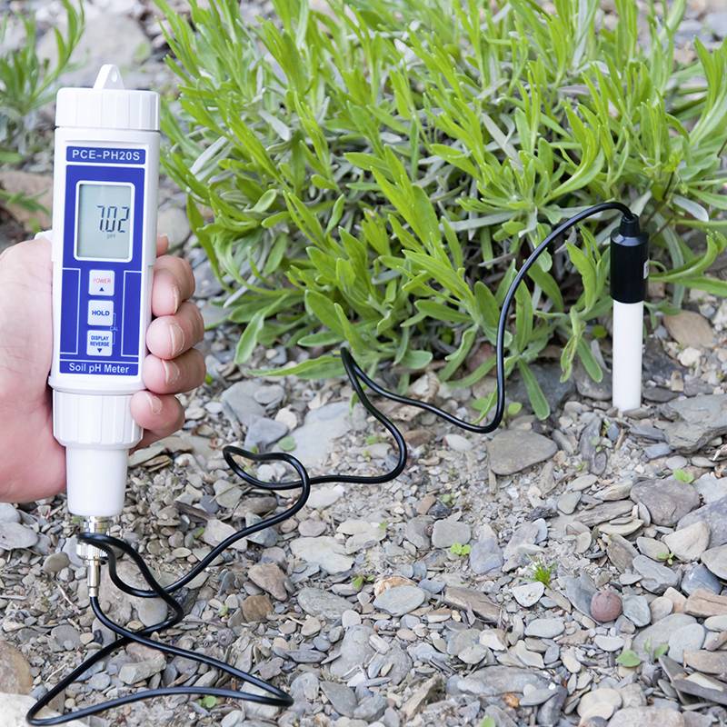 Une main tient un dispositif de mesure du pH du sol, affichant la valeur '7,02'. L'appareil est enfoncé dans un sol pierreux avec des plantes.