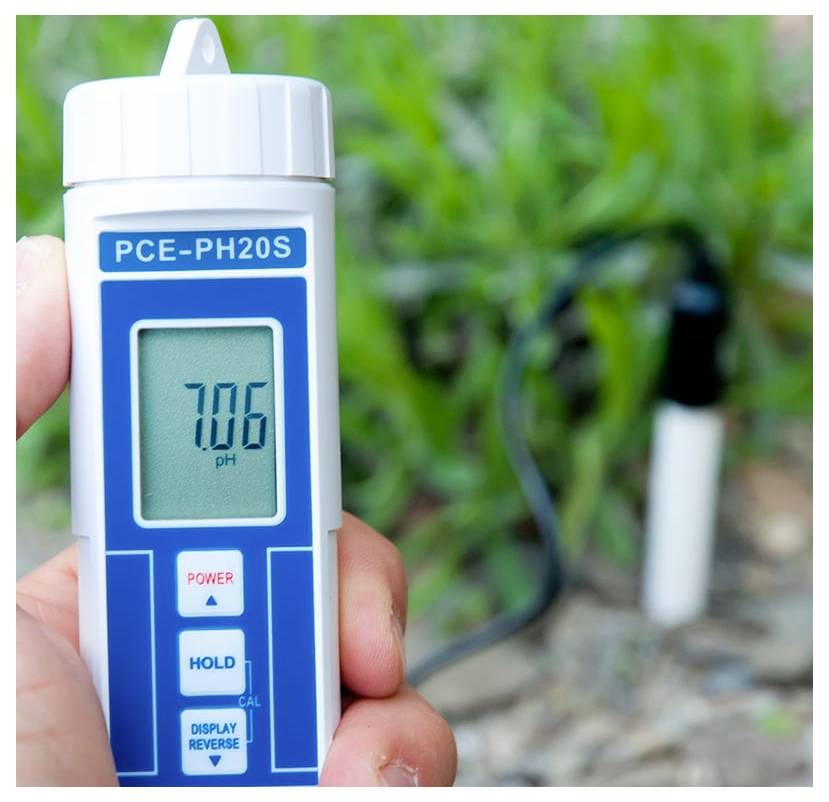 Mesure de pH avec le lecteur portable PCE-PH20S affichant 7,06, mesurant le sol dans un jardin avec une sonde en arrière-plan.