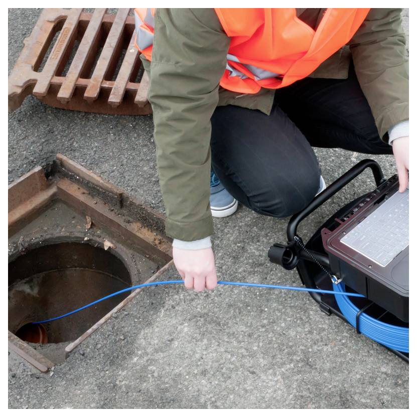 Ouvrier en gilet de sécurité orange abaisse un câble bleu dans un regard ouvert à côté d'un puisard de rue, utilisant un équipement d'inspection.