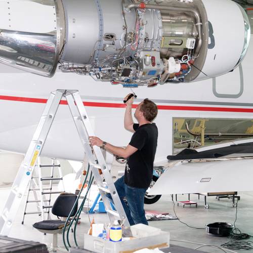 Un technicien est perché sur une échelle et examine un moteur d'avion à l'aide d'une lampe de poche dans un hangar.