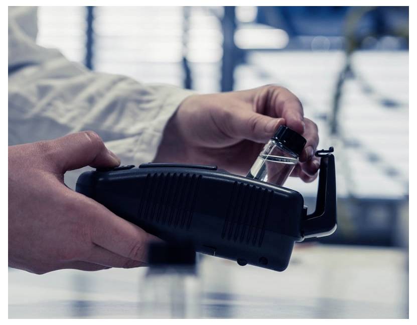Les mains insèrent une petite fiole dans un appareil portatif noir, probablement un scanner d'échantillon ou un instrument de laboratoire, dans un environnement de laboratoire flou.