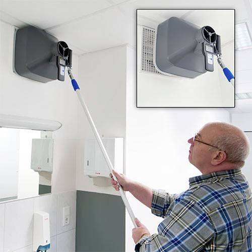Un homme mesure le flux d'air à une ouverture de ventilation dans un bureau ou un laboratoire à l'aide d'un appareil fixé au plafond.