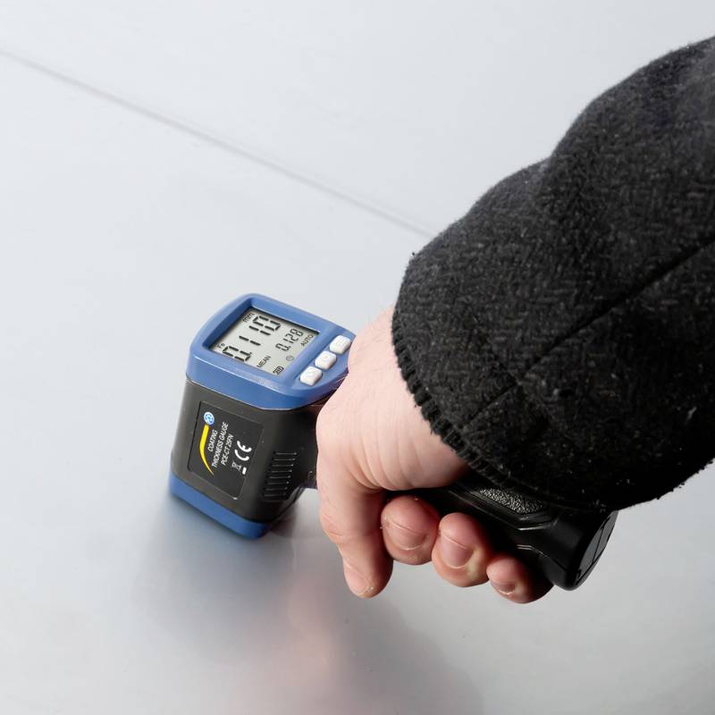 Un appareil portable mesure l'épaisseur d'une surface. L'affichage indique '1.18'. Une main tient l'appareil contre une surface grise.