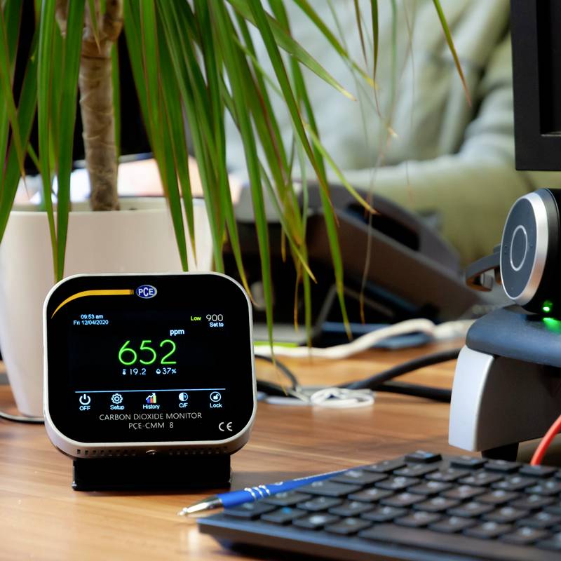 Une table avec un moniteur de CO2 affichant 652 ppm, placé à côté d'un écran d'ordinateur. Une plante se trouve en arrière-plan.
