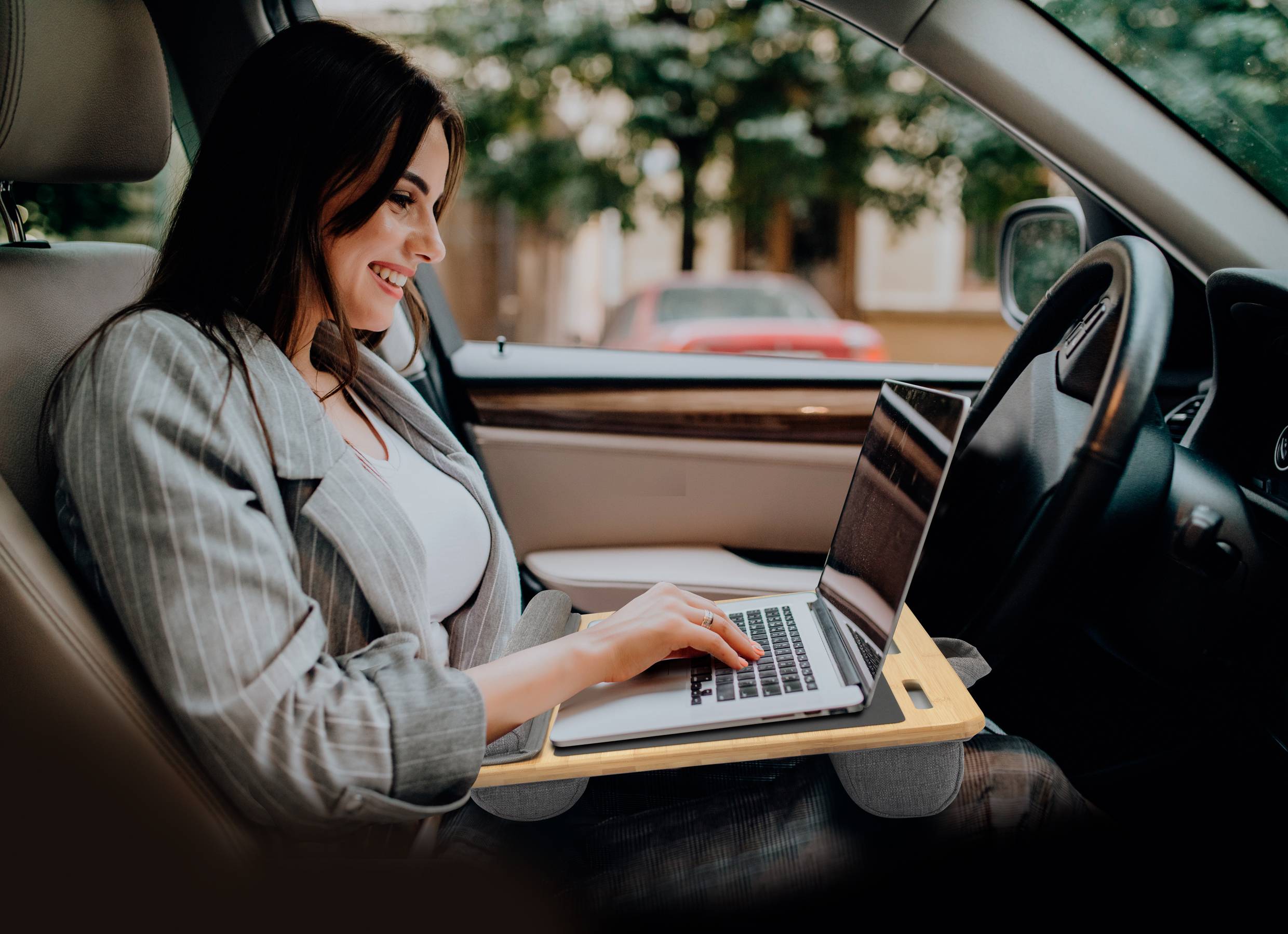 Une femme sourit tout en travaillant sur un ordinateur portable posé sur une tablette dans une voiture, garée avec des arbres en arrière-plan.