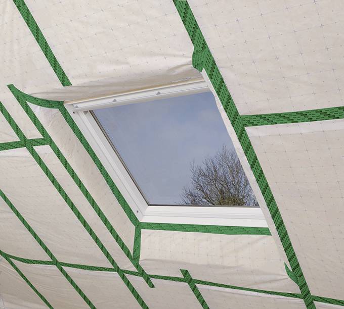 Fenêtre intégrée dans un plafond avec des joints d'étanchéité verts et un film isolant, offrant une vue sur l'extérieur. Un ciel bleu clair est visible.