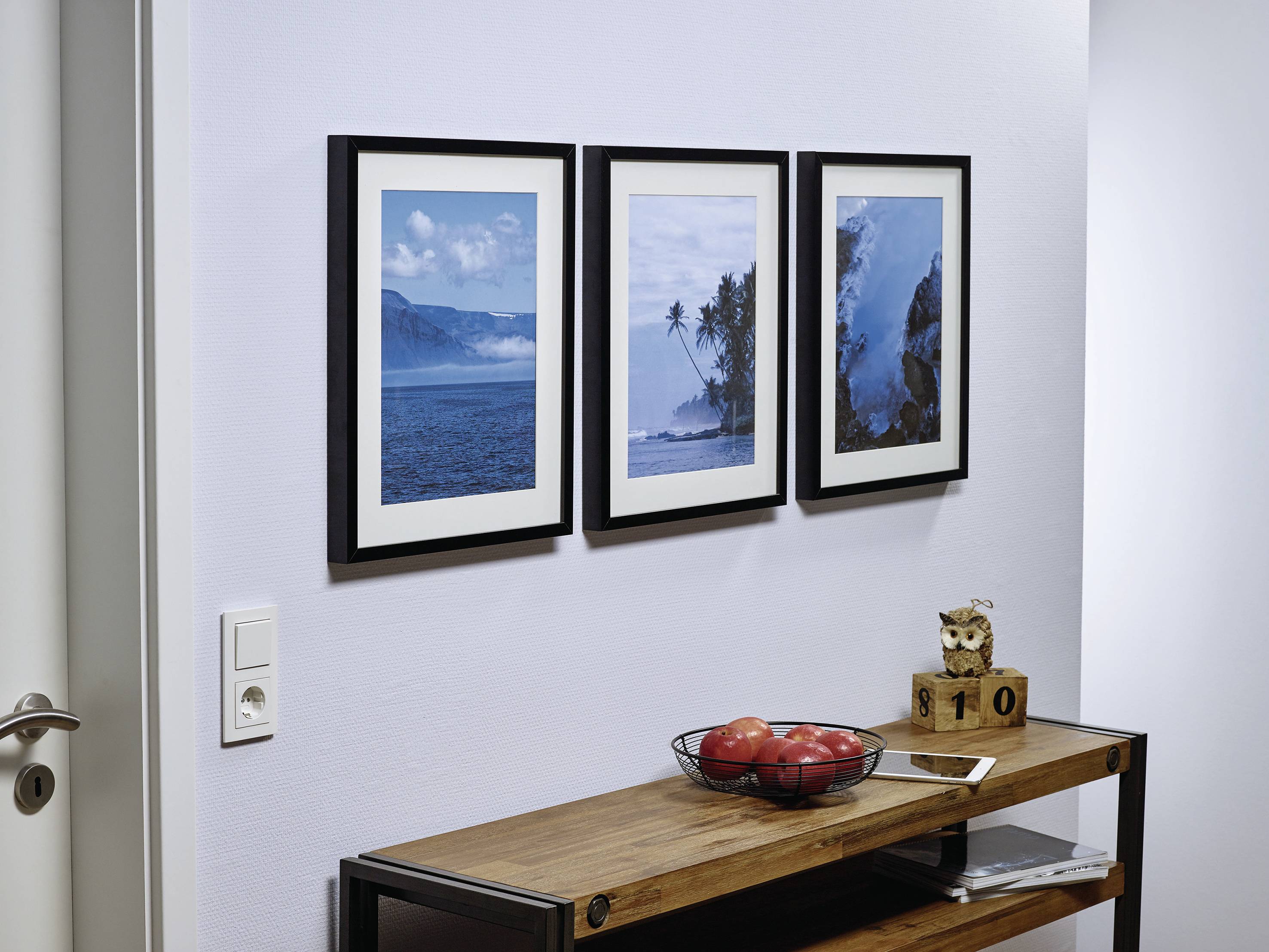 Trois tableaux de paysage encadrés sont suspendus au-dessus d'une table en bois portant un bol de pommes et une chouette décorative.