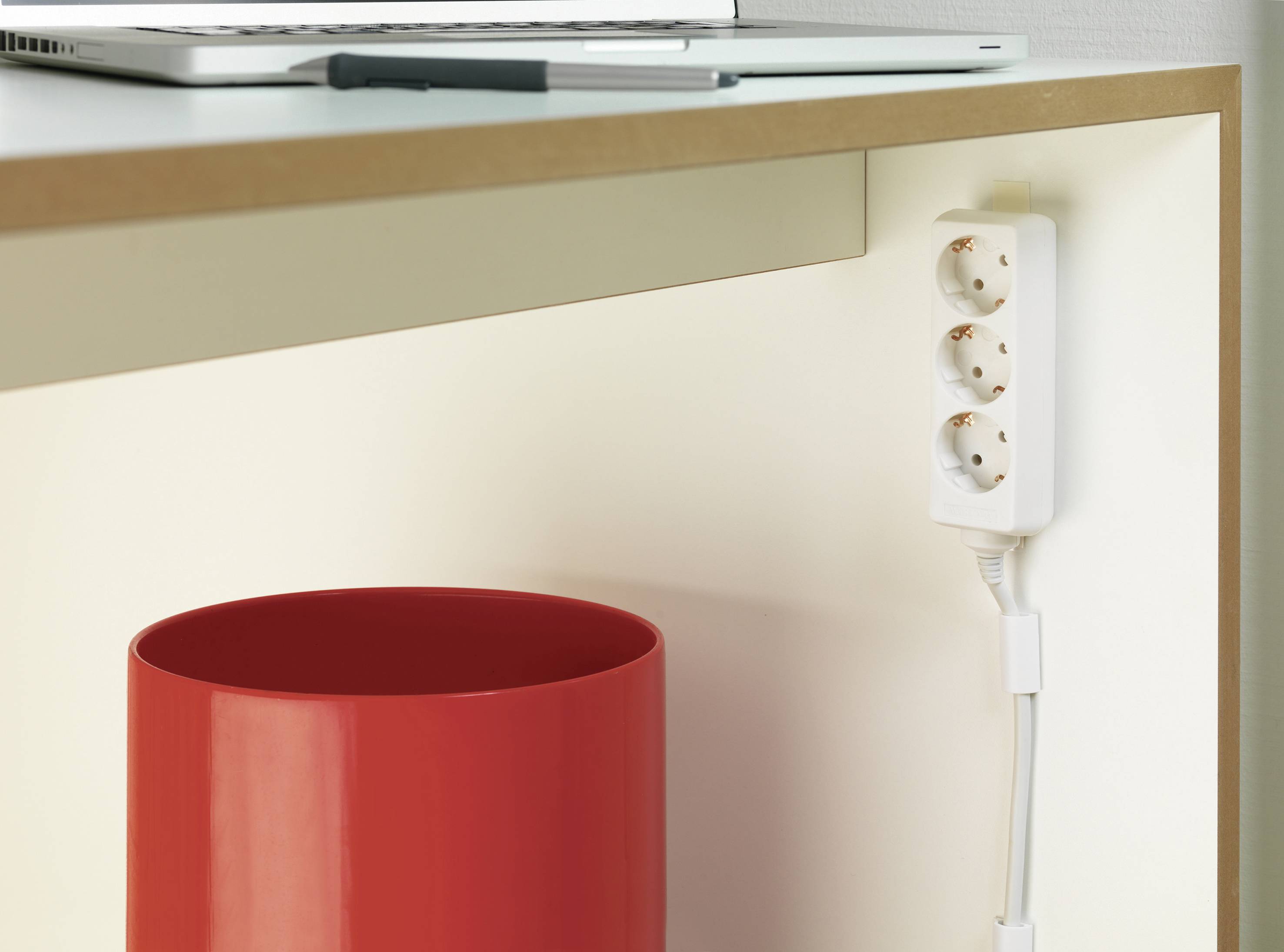 Une multiprise extensible est suspendue sous un bureau, à côté d'une poubelle rouge. Un ordinateur portable et un stylo sont visibles sur le dessus.