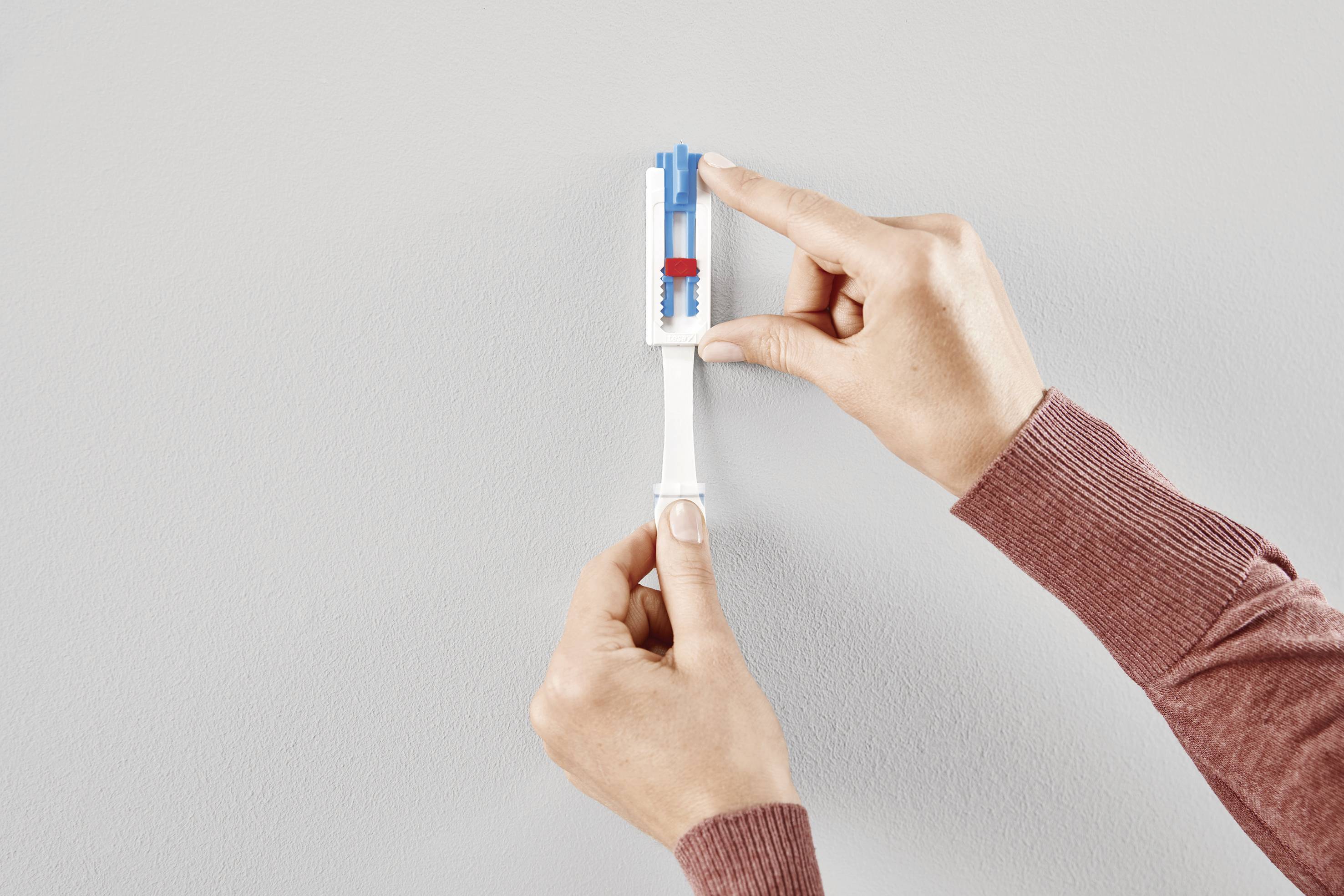 Deux mains installent un objet bleu et rouge sur un support blanc fixé au mur.