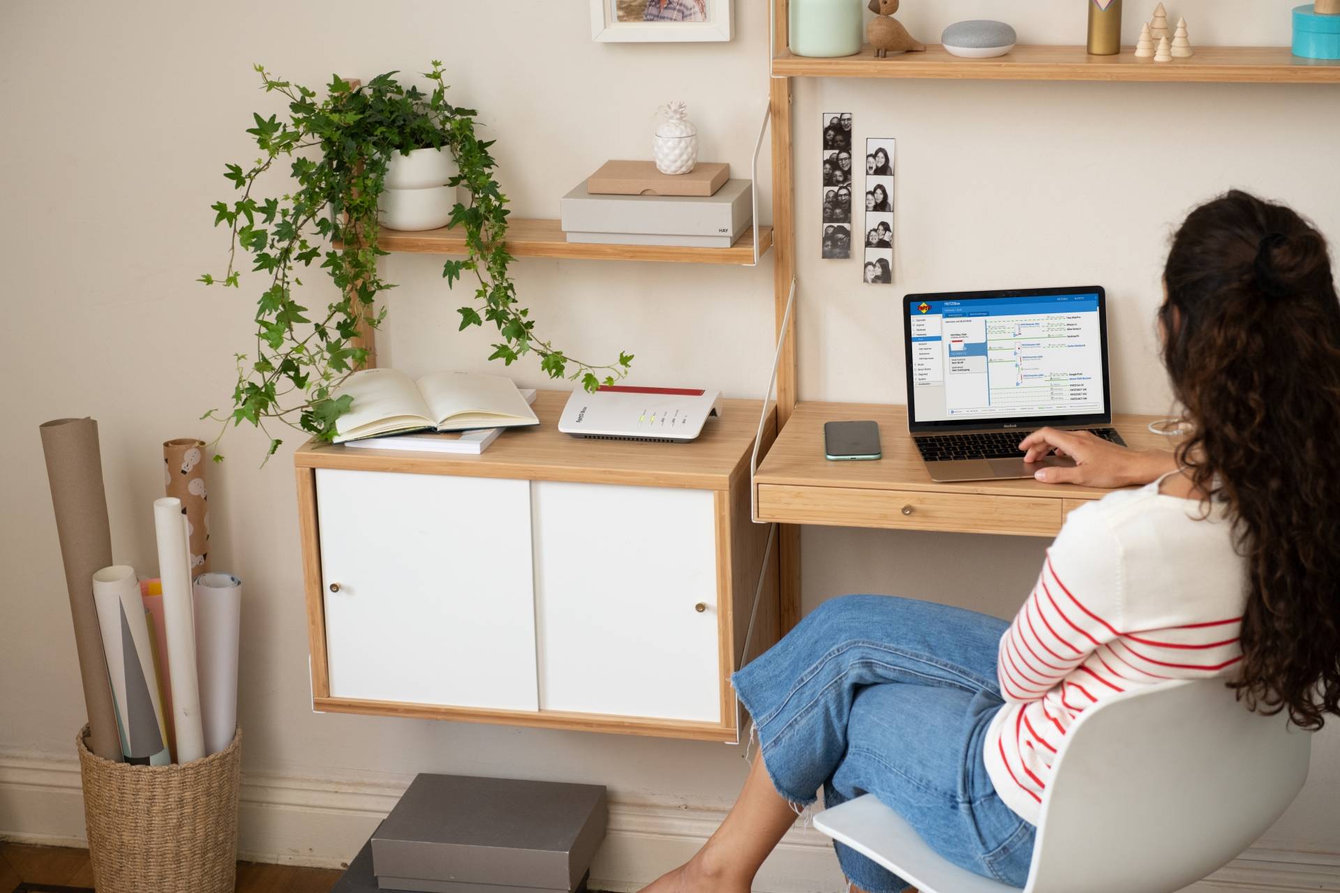 Une femme est assise à un bureau avec un ordinateur portable. Elle travaille dans une pièce chaleureuse avec des plantes et des étagères en arrière-plan.