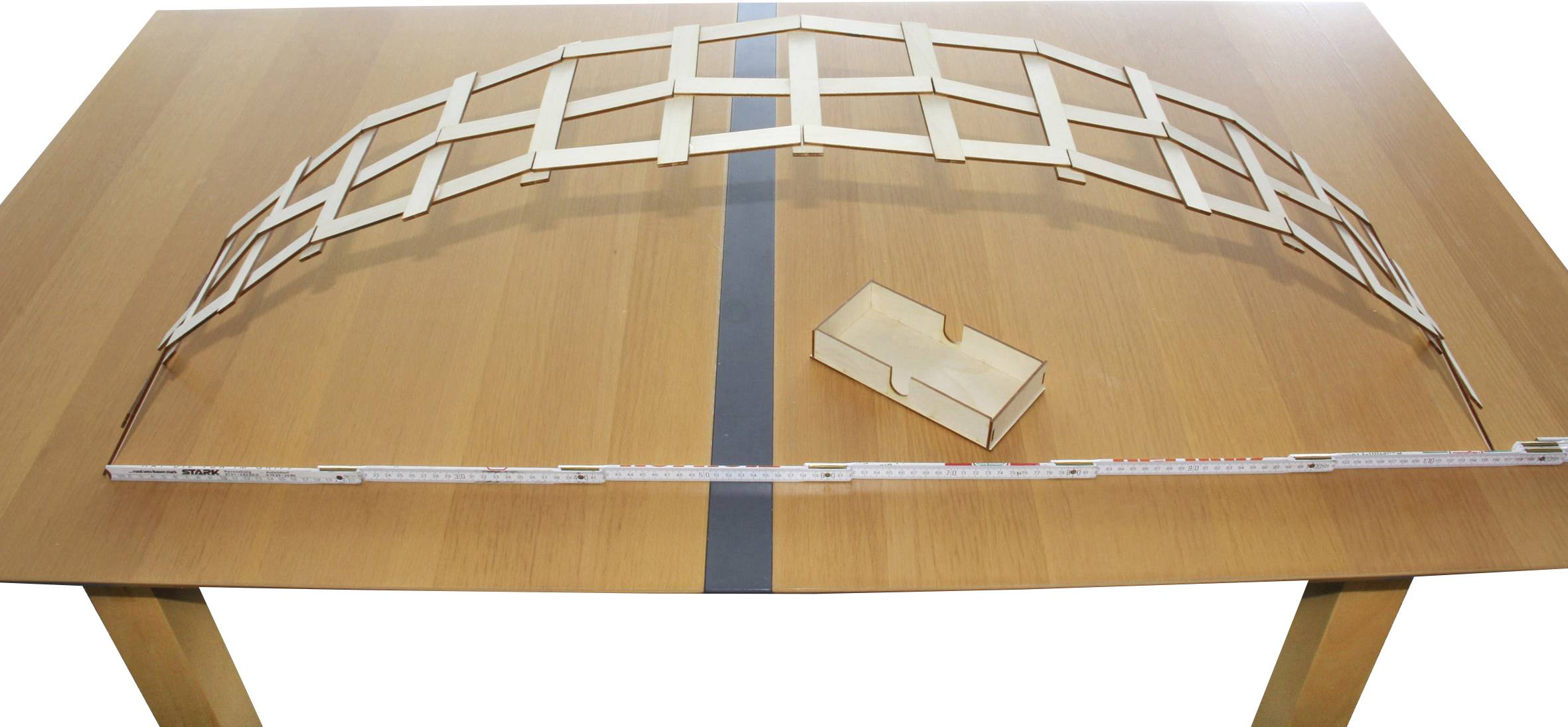 Un modèle de voûte en bois composé de barres plates repose sur une table. À côté se trouve une petite boîte en bois. Un mètre ruban s'étend le long de l'arc.