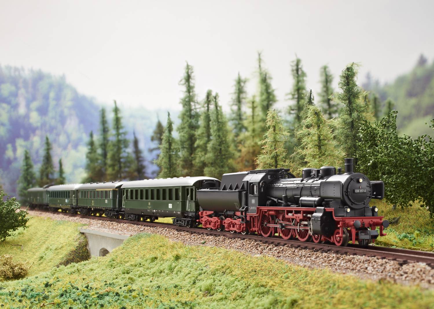 Un train miniature traverse un paysage avec des arbres et des collines, composé d'une locomotive à vapeur et de wagons verts.