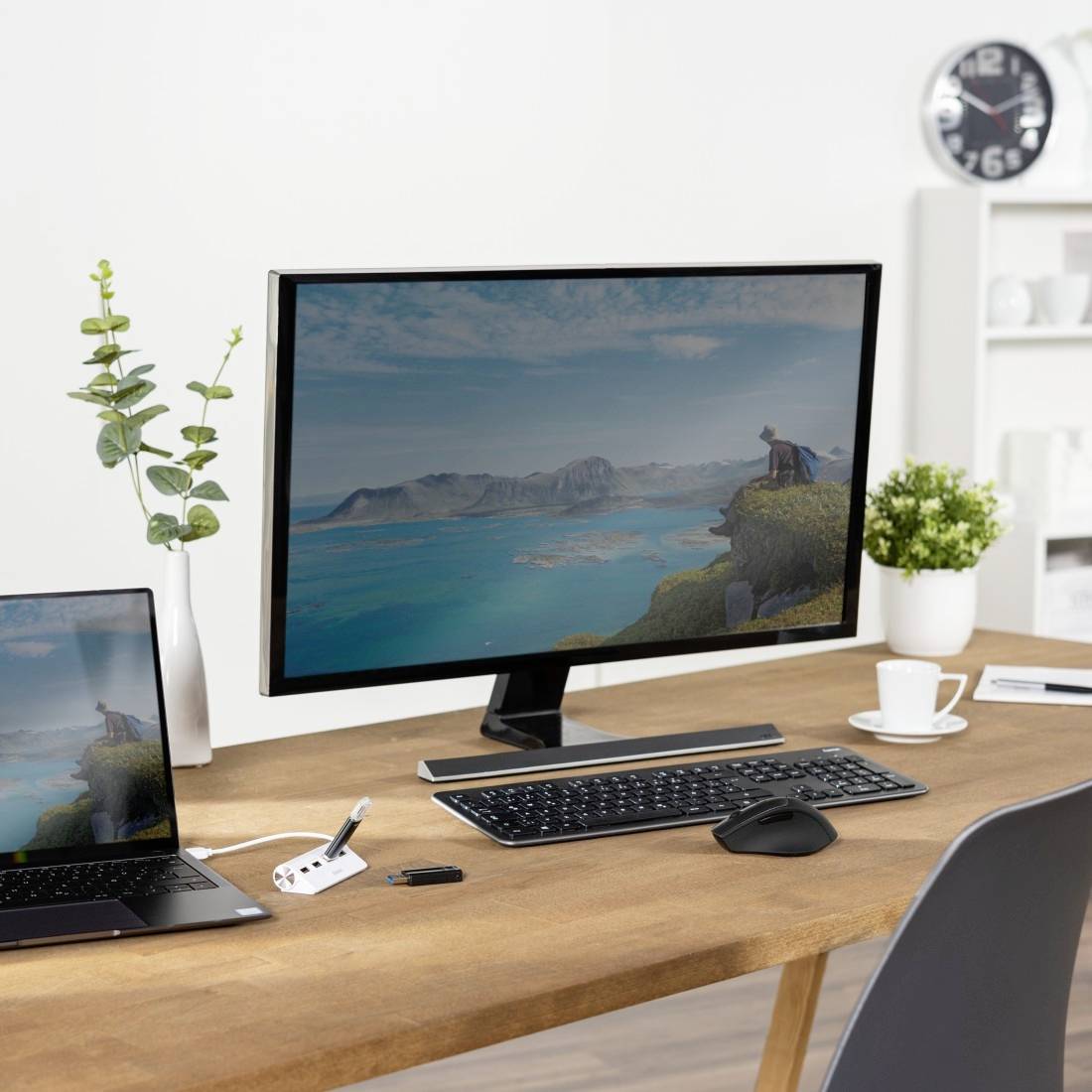 Un bureau avec un écran d'ordinateur affichant un paysage, entouré de plantes, d'un ordinateur portable, d'un clavier, d'une souris et d'une tasse de café.