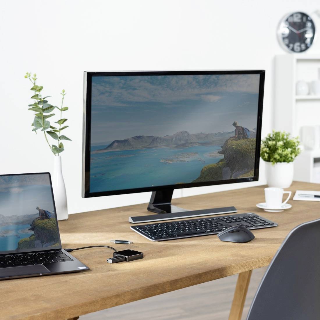 Un bureau avec un ordinateur portable et un écran affichant un paysage. Un clavier et une souris sont posés à côté. En arrière-plan, on aperçoit une horloge et des étagères.
