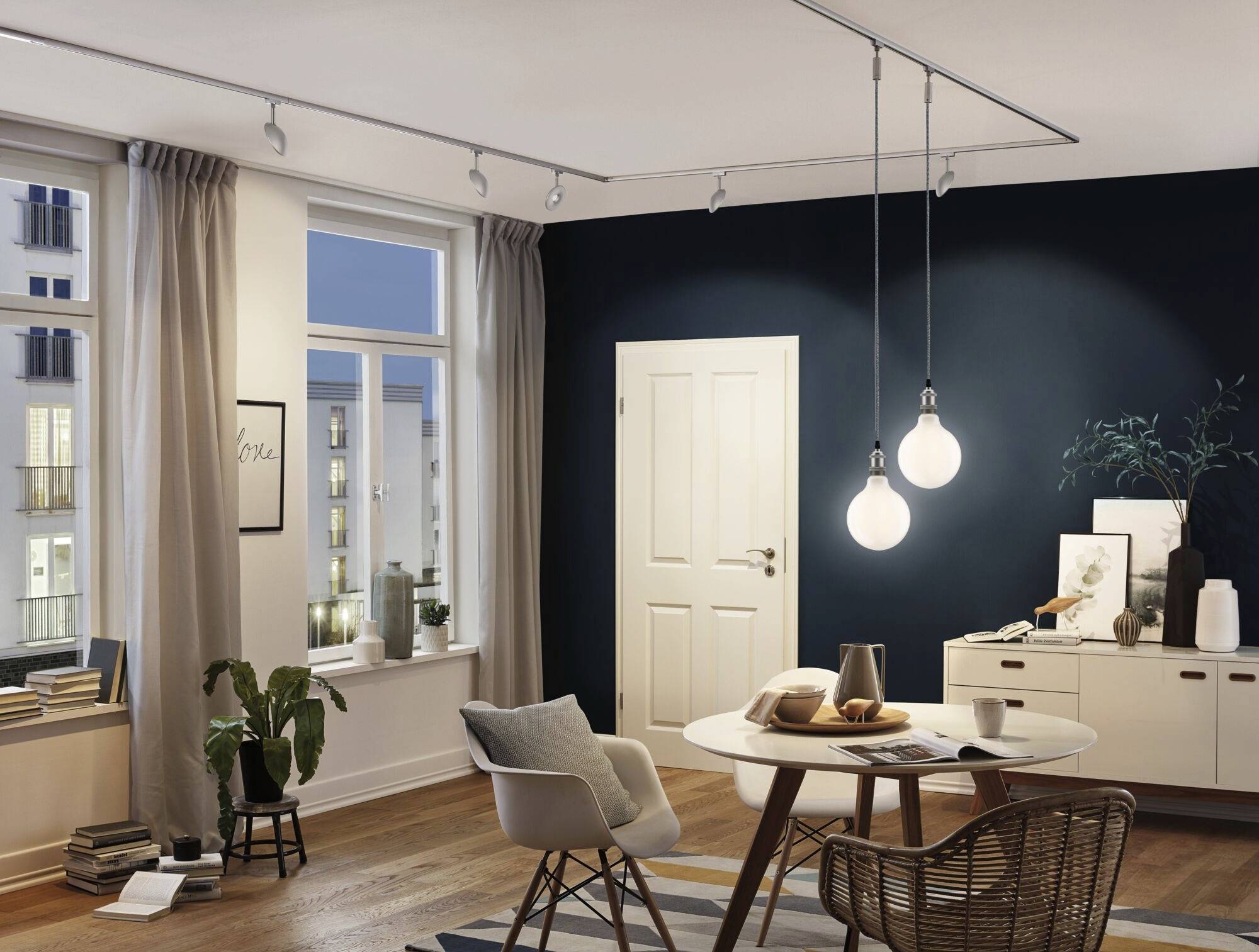 Un salon accueillant avec un sol en bois, une table ronde et des suspensions. Les fenêtres donnent sur des bâtiments urbains à la tombée du jour.