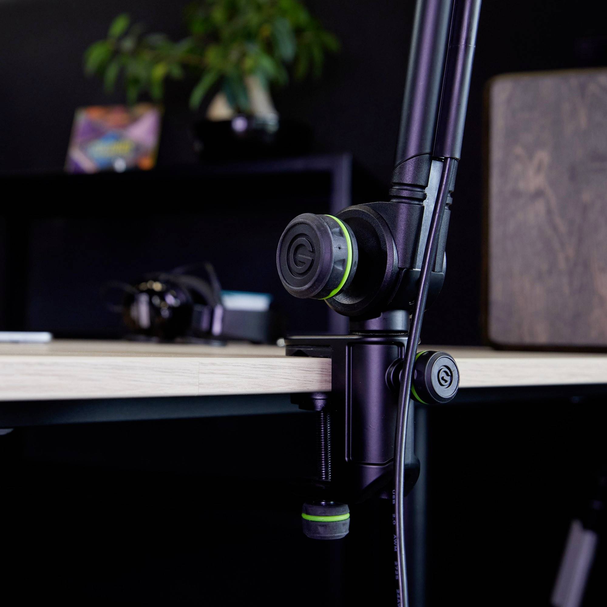 Un bras de microphone est fixé à une table. En arrière-plan, des écouteurs et une plante sont visibles de manière floue.