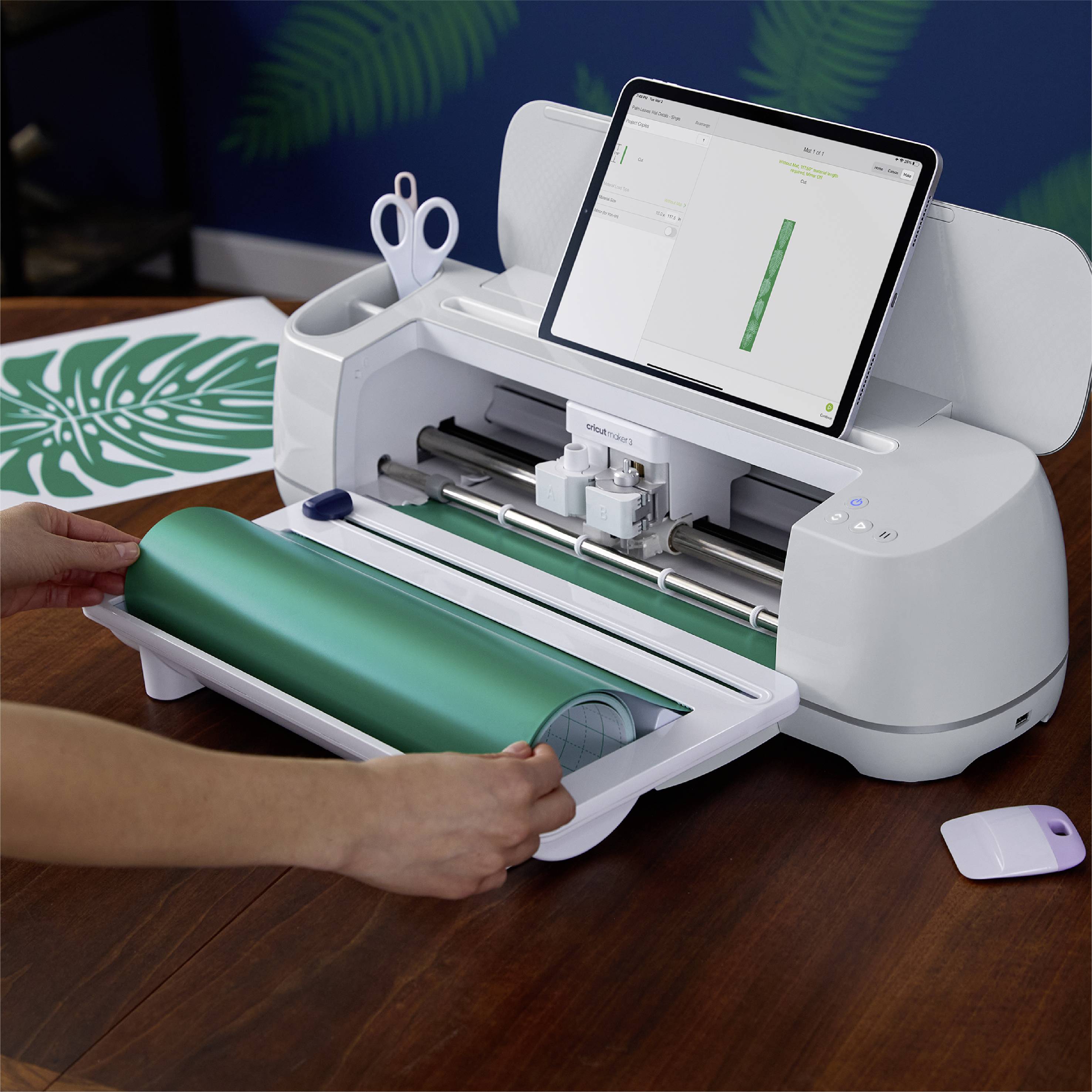 Un traceur de découpe sur une table coupe du vinyle vert. Une tablette affiche un design de feuille. Une main tient un rouleau de vinyle.