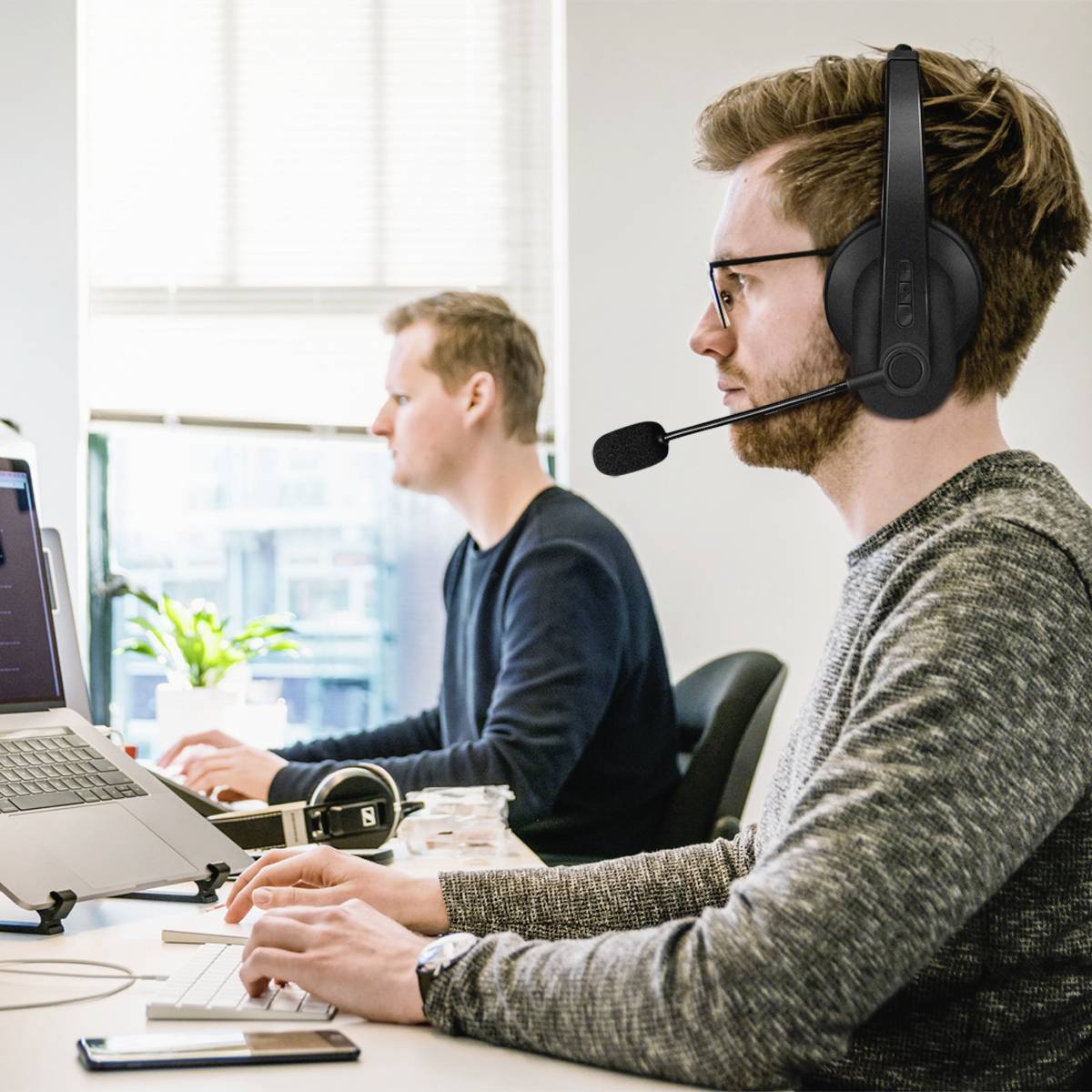 Deux hommes travaillent sur des ordinateurs dans un bureau. L'homme à l'avant-plan porte un casque avec microphone. À l'arrière-plan, une plante est située près de la fenêtre.